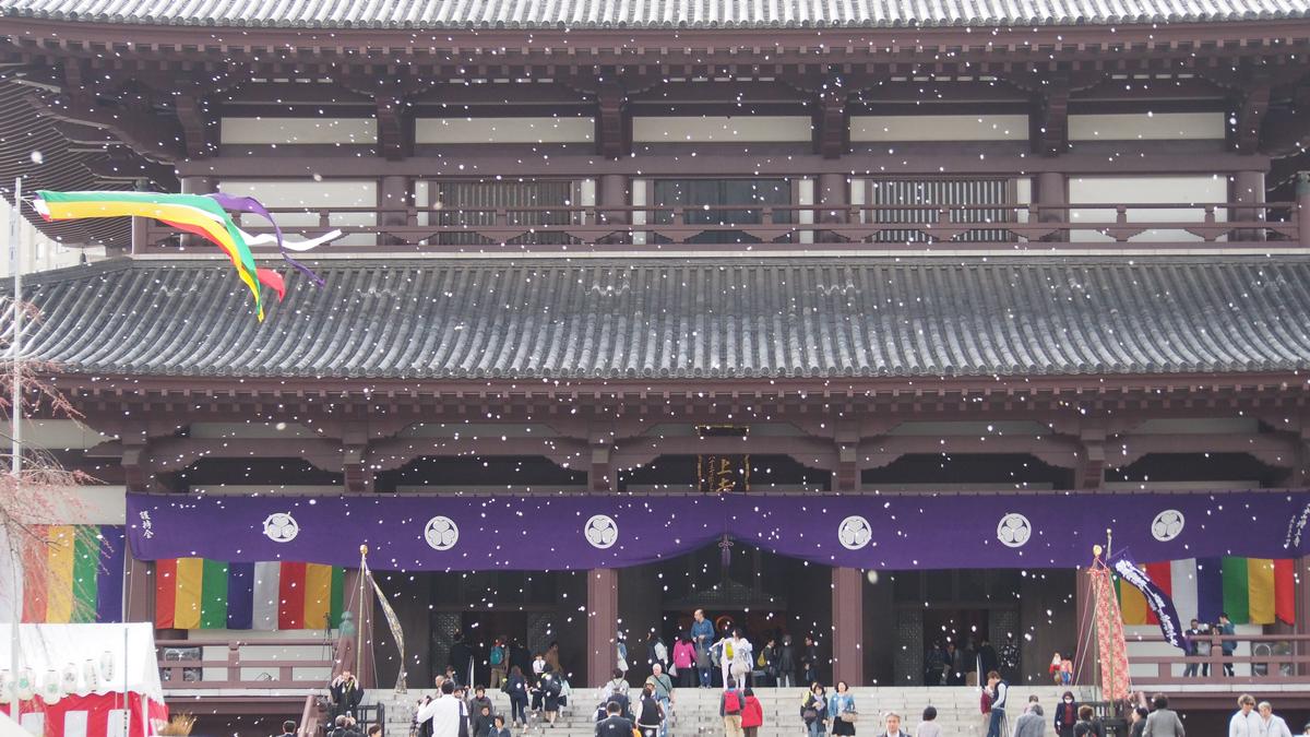 White confetti falls over a large Japanese temple building adorned with colorful banners and a crowd of people.