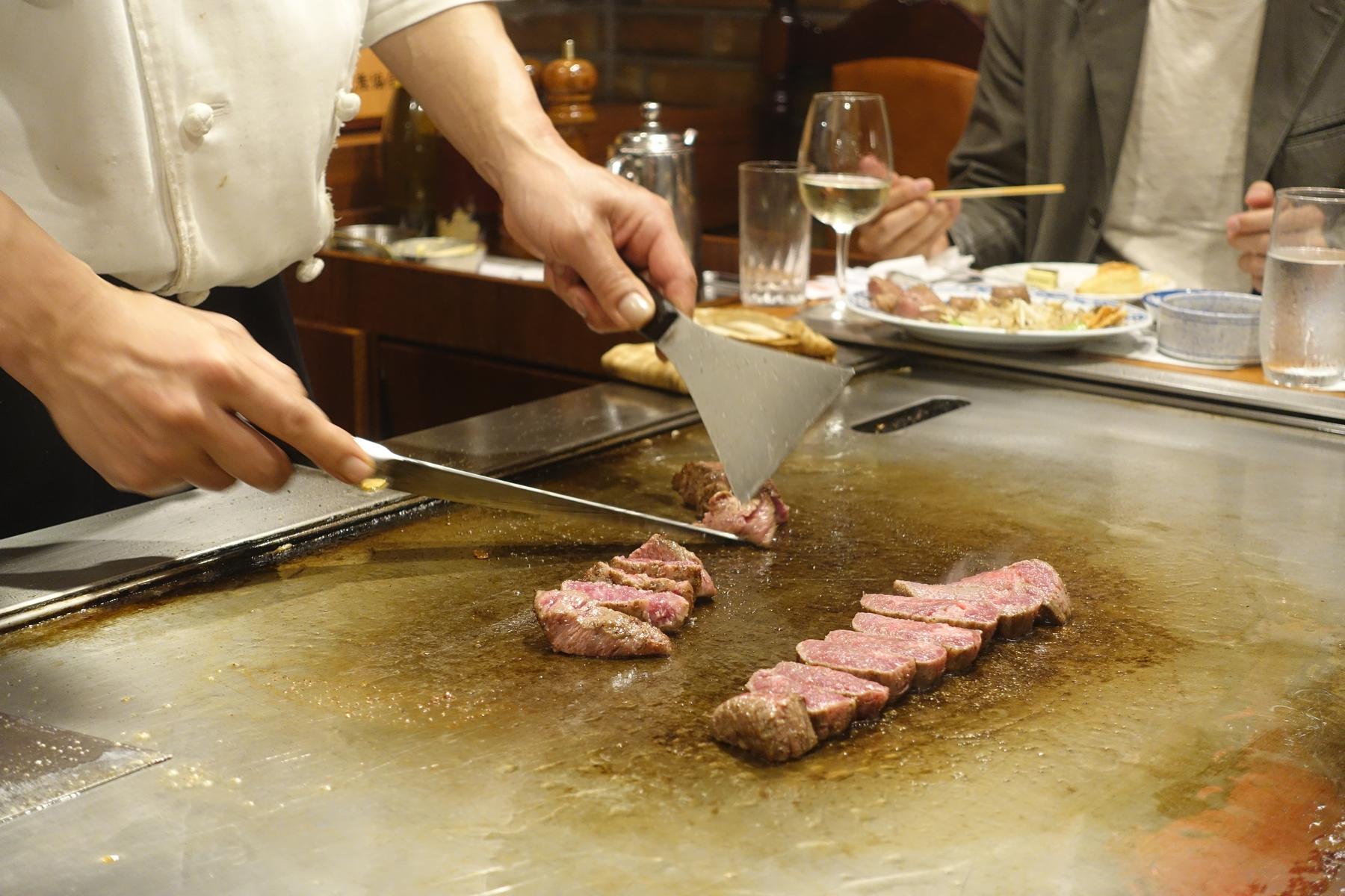 A chef cooks sliced steak on a teppanyaki grill as a diner watches.