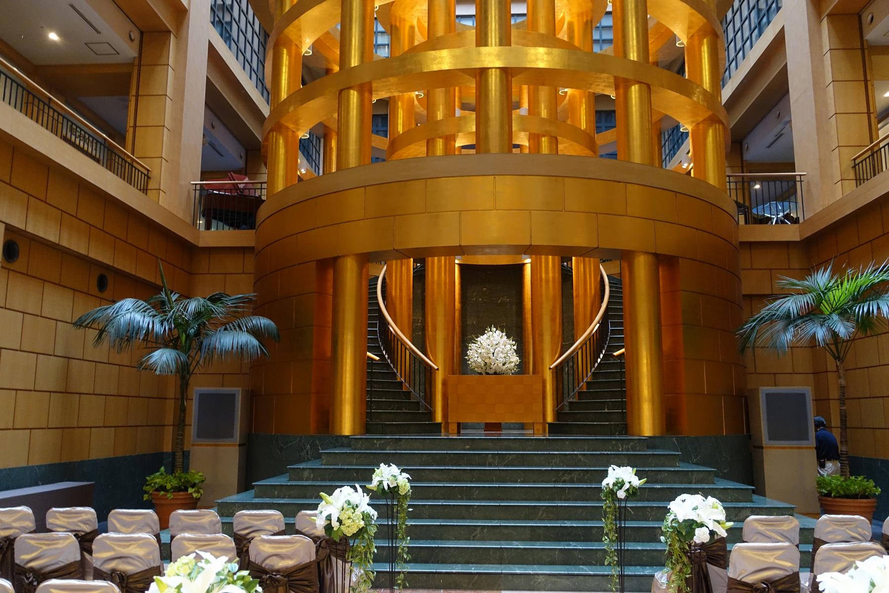 A grand hall featuring a prominent golden cylindrical structure, marble staircase, and rows of chairs for an event.