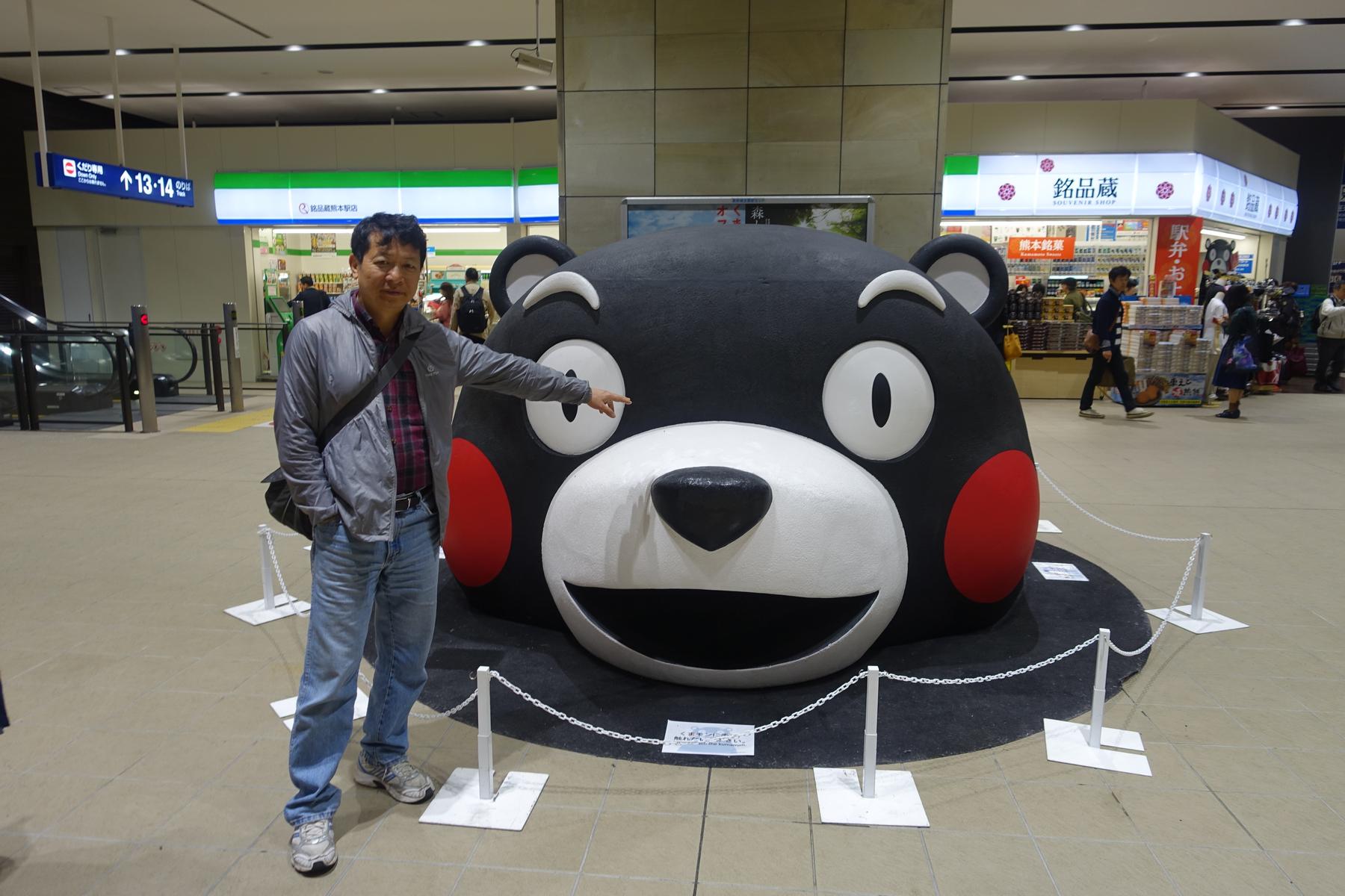A man points to a large Kumamon mascot in a Japanese station.