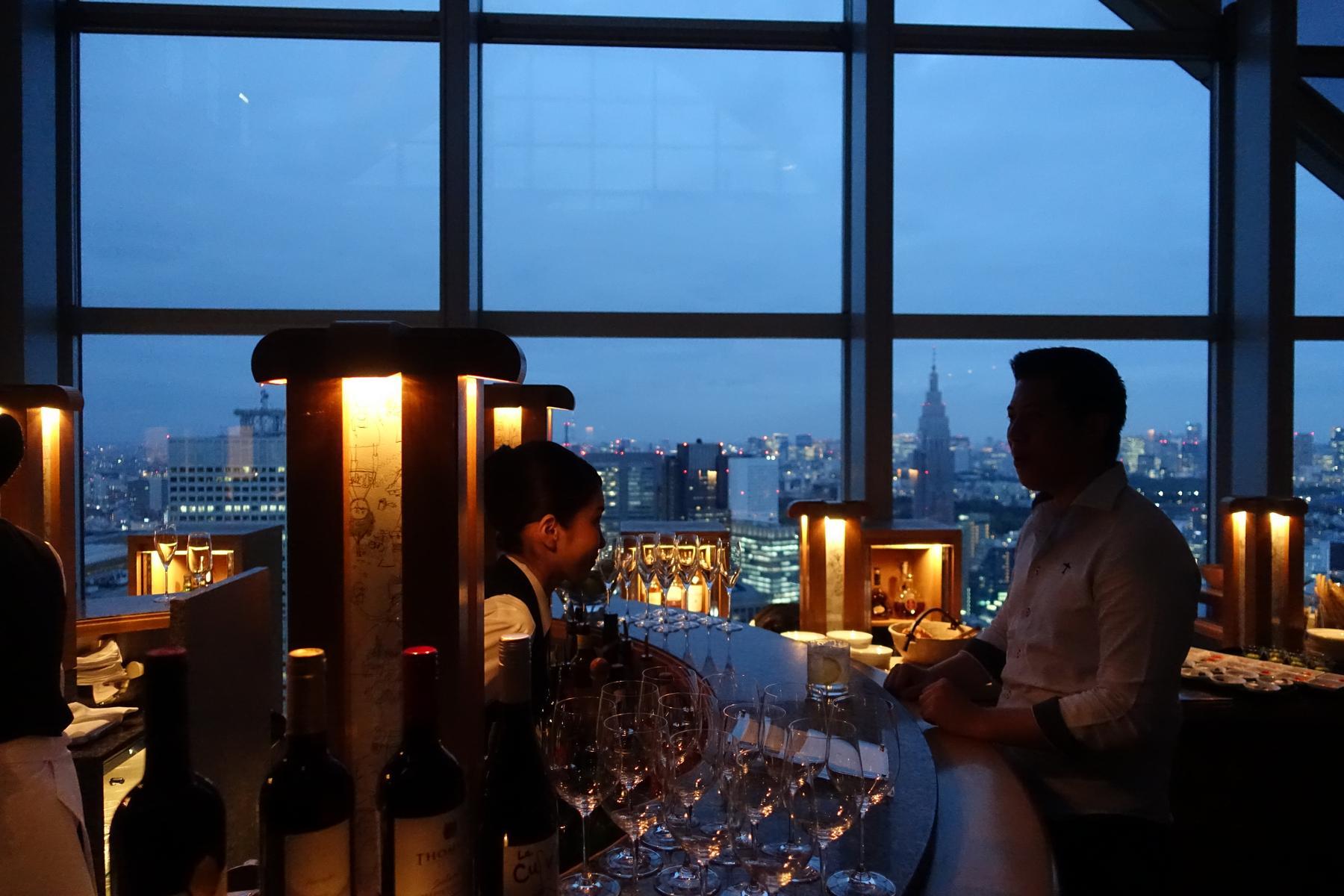 A bartender and patron at a high-rise bar with a twilight city skyline view.