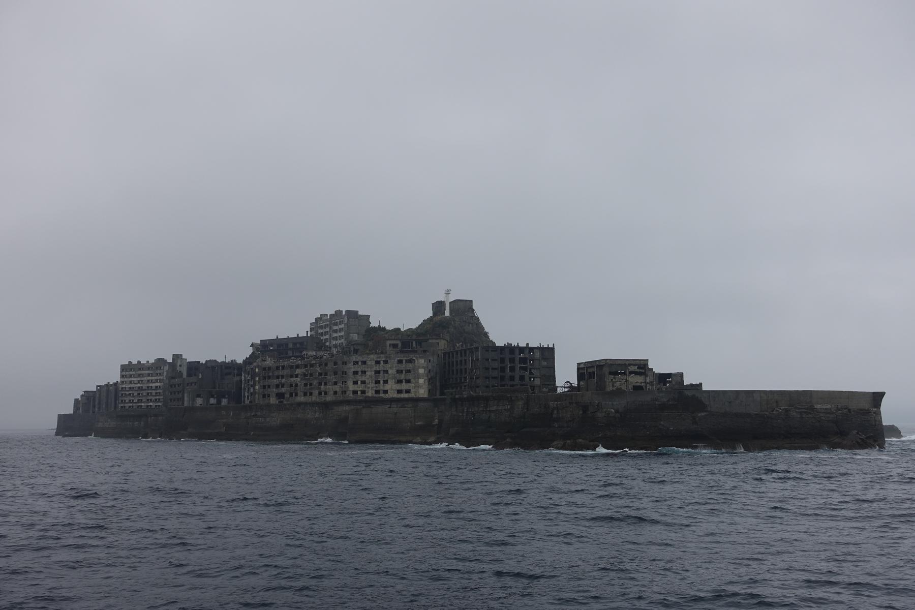 Hashima Island (Gunkanjima) with its abandoned concrete buildings rises from the choppy sea under a grey sky.
