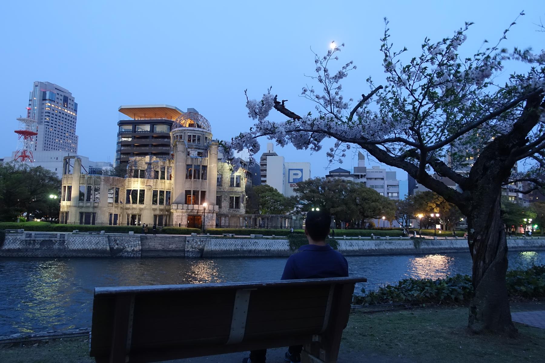 A person sits on a park bench facing the Hiroshima Peace Memorial (Atomic Bomb Dome) and a river, with cherry blossoms in the foreground at dusk.