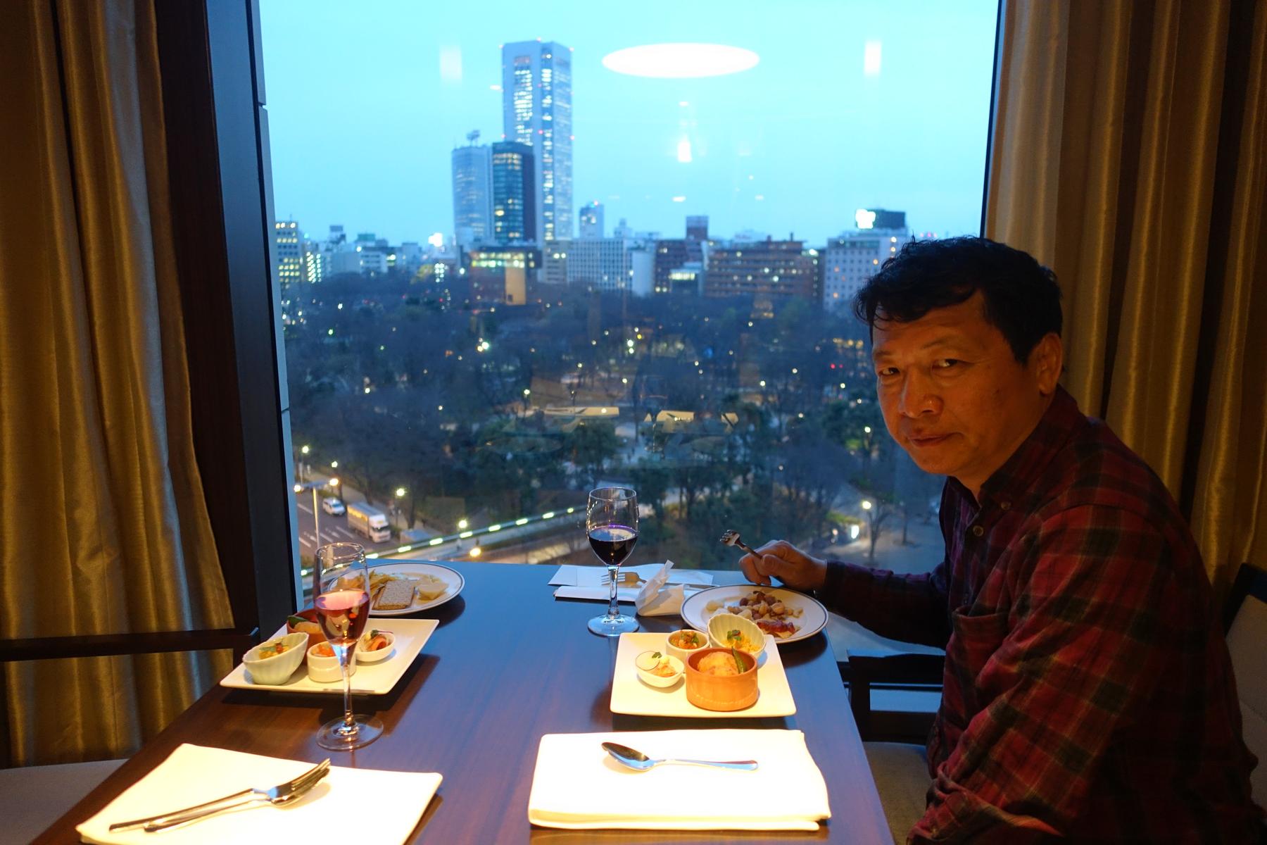 Man dining at a table by a large window overlooking a city at dusk.
