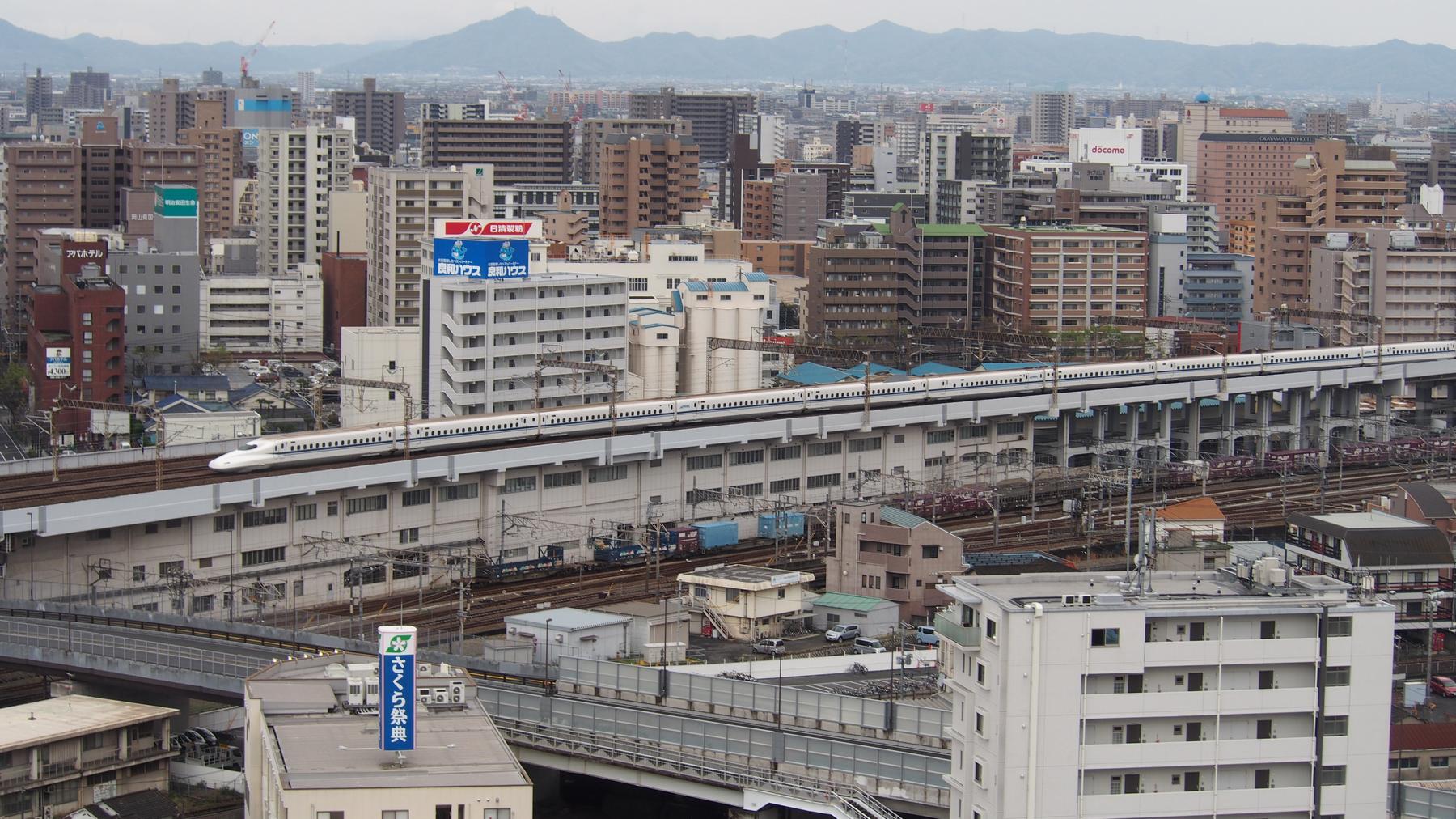 A white Shinkansen bullet train rides an elevated track through a dense Japanese city.