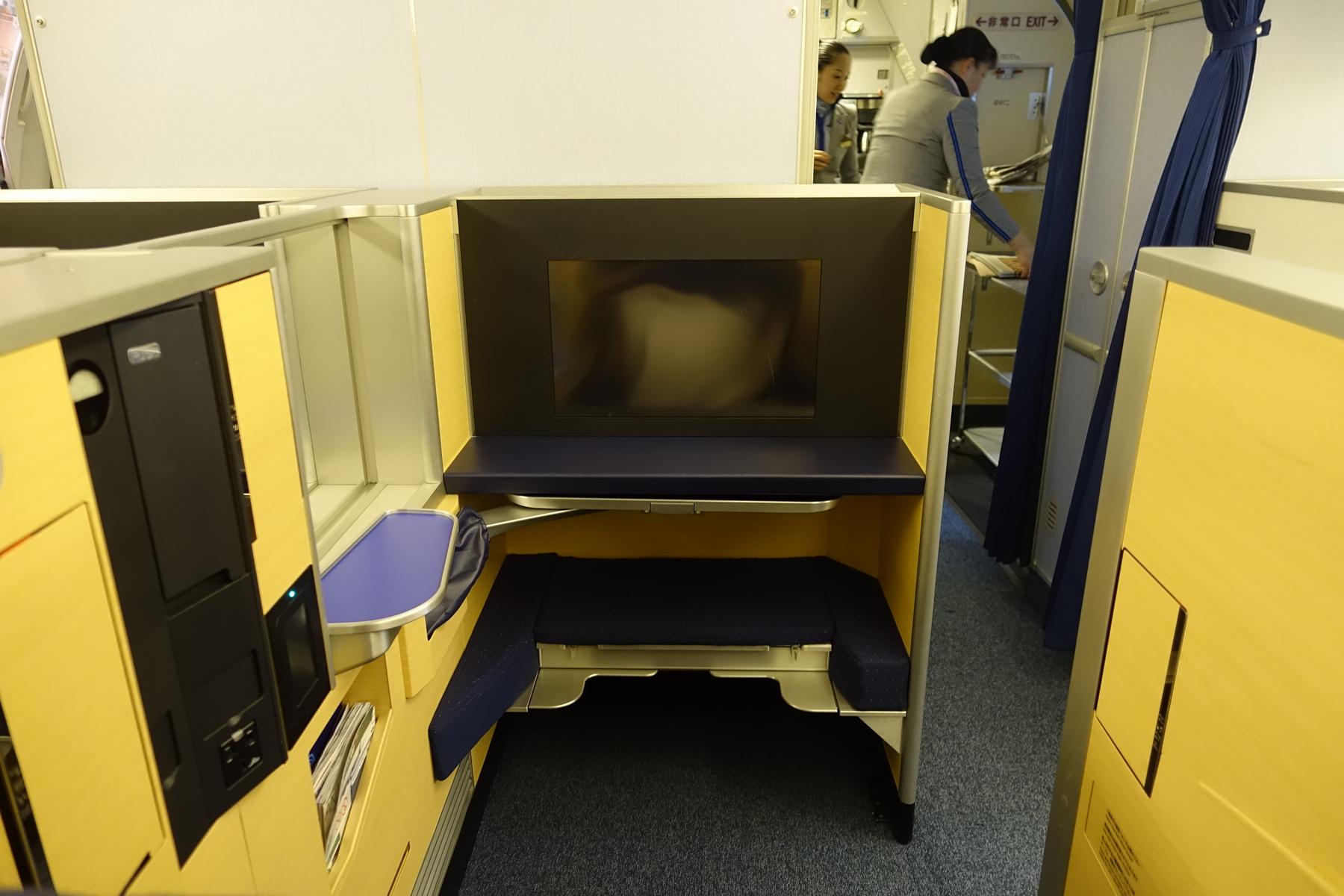 An empty business class airplane seat with a large personal screen, as flight attendants work in the background.