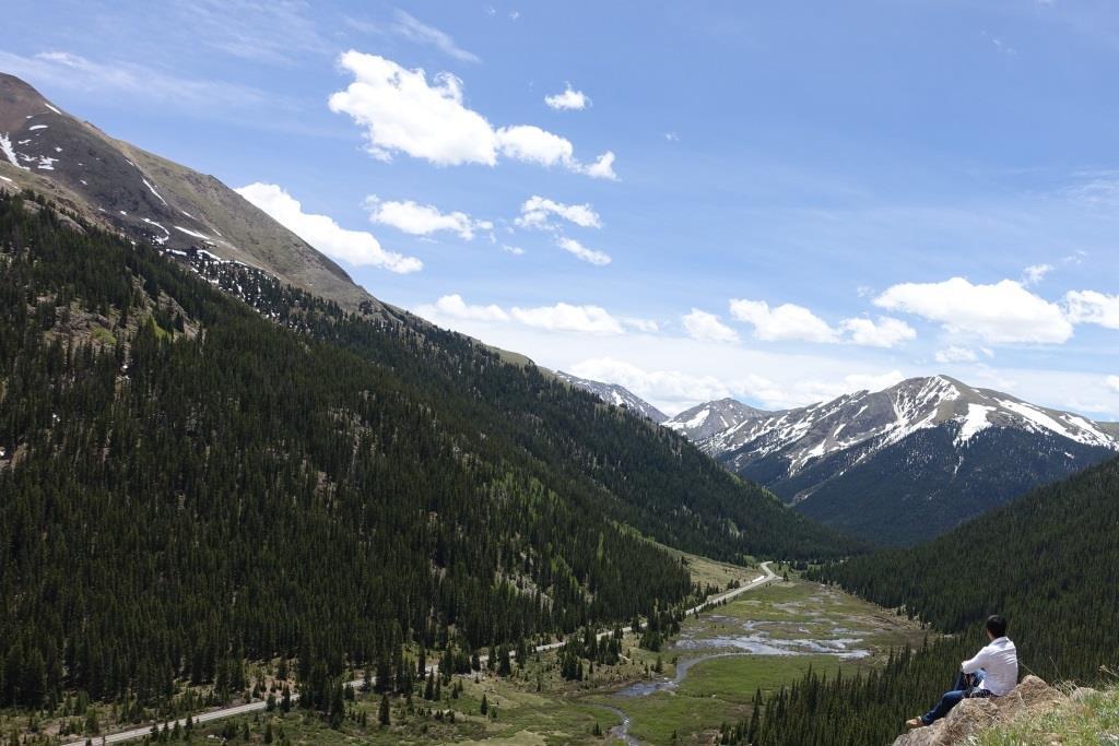 A person sits on a rock overlooking a mountain valley with a winding road, dense forests, and snowy peaks.