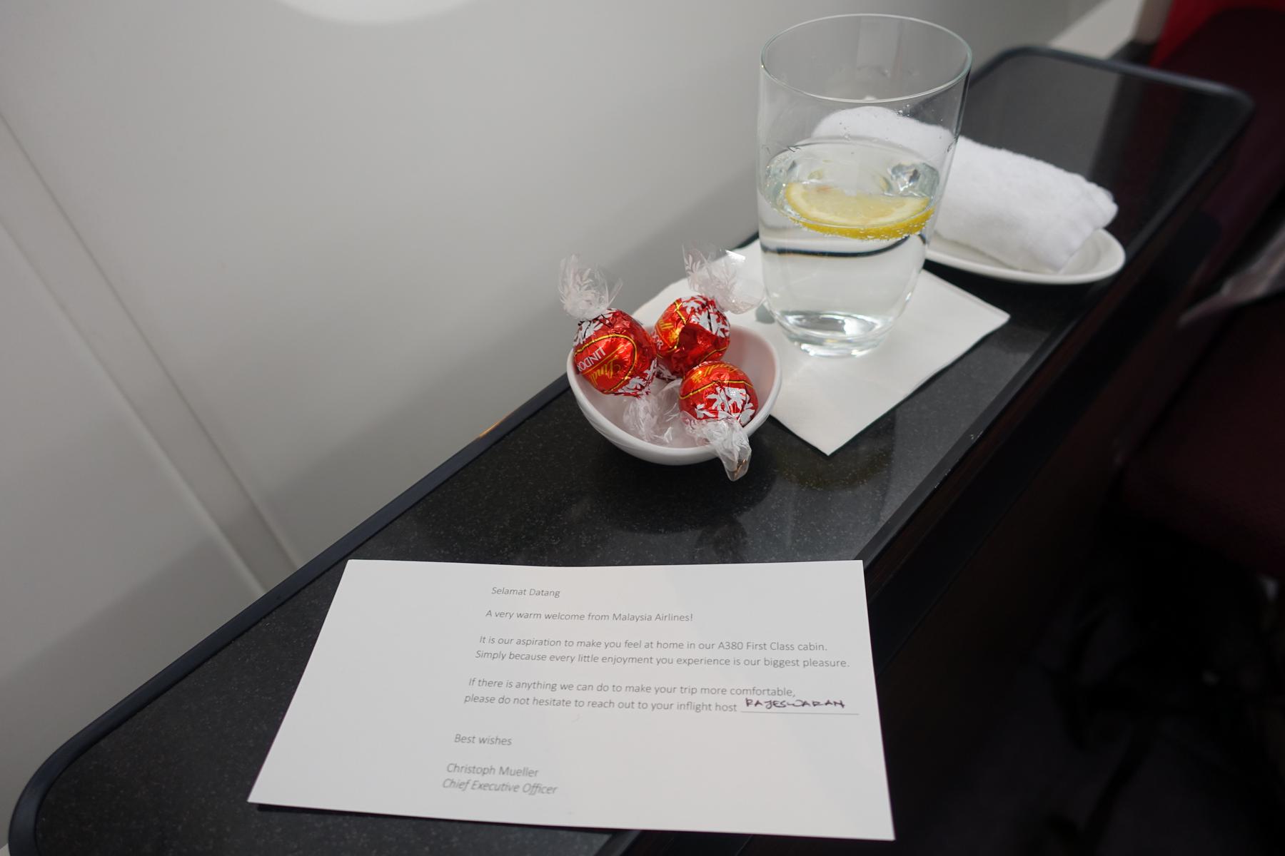 A welcome letter, a glass of water with lemon, and Lindor chocolates on a Malaysia Airlines First Class tray table.