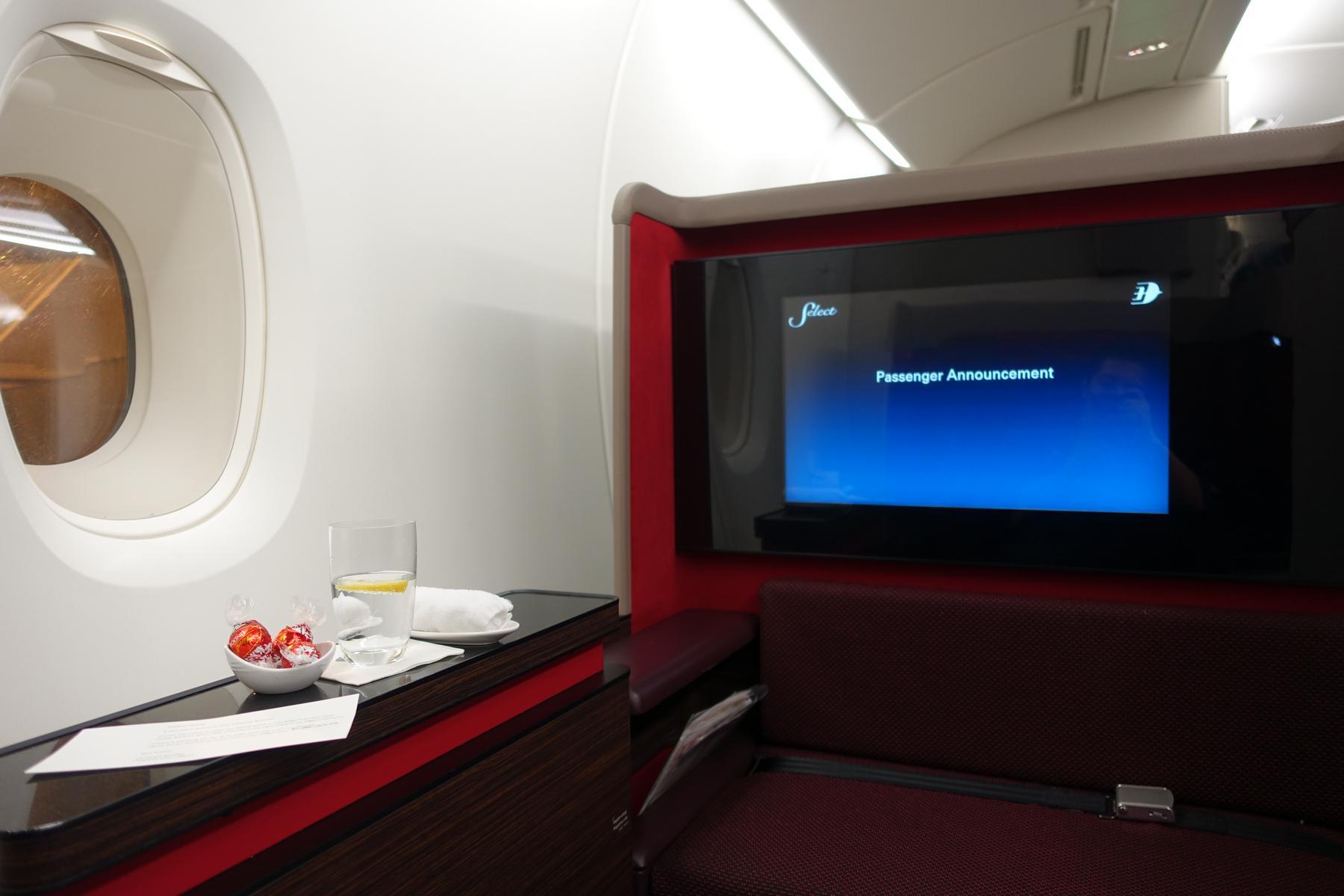 A Malaysia Airlines A380 First Class suite with a window, personal screen displaying an announcement, and a side table with a glass of water and chocolates.
