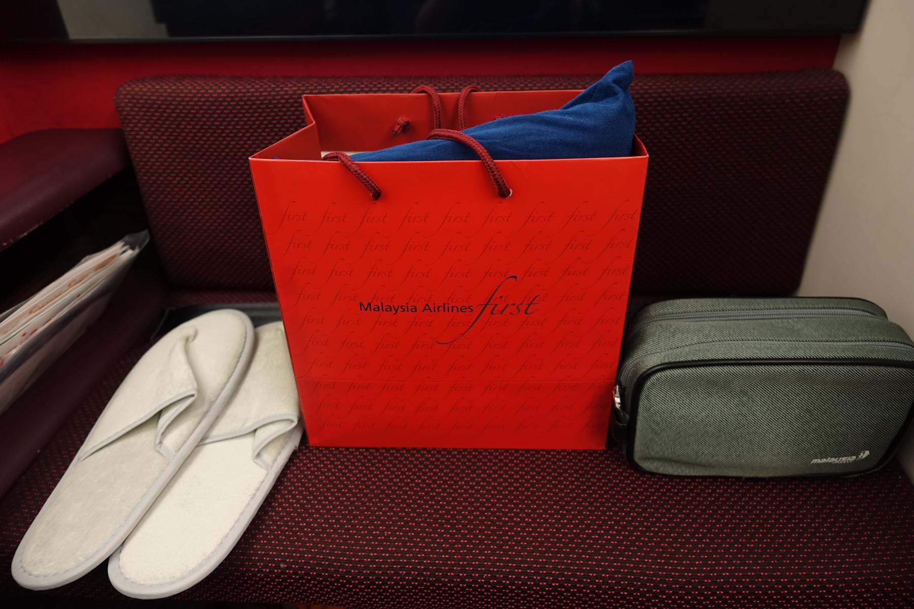A red Malaysia Airlines First Class gift bag, slippers, and amenity kit sit on a cabin seat.