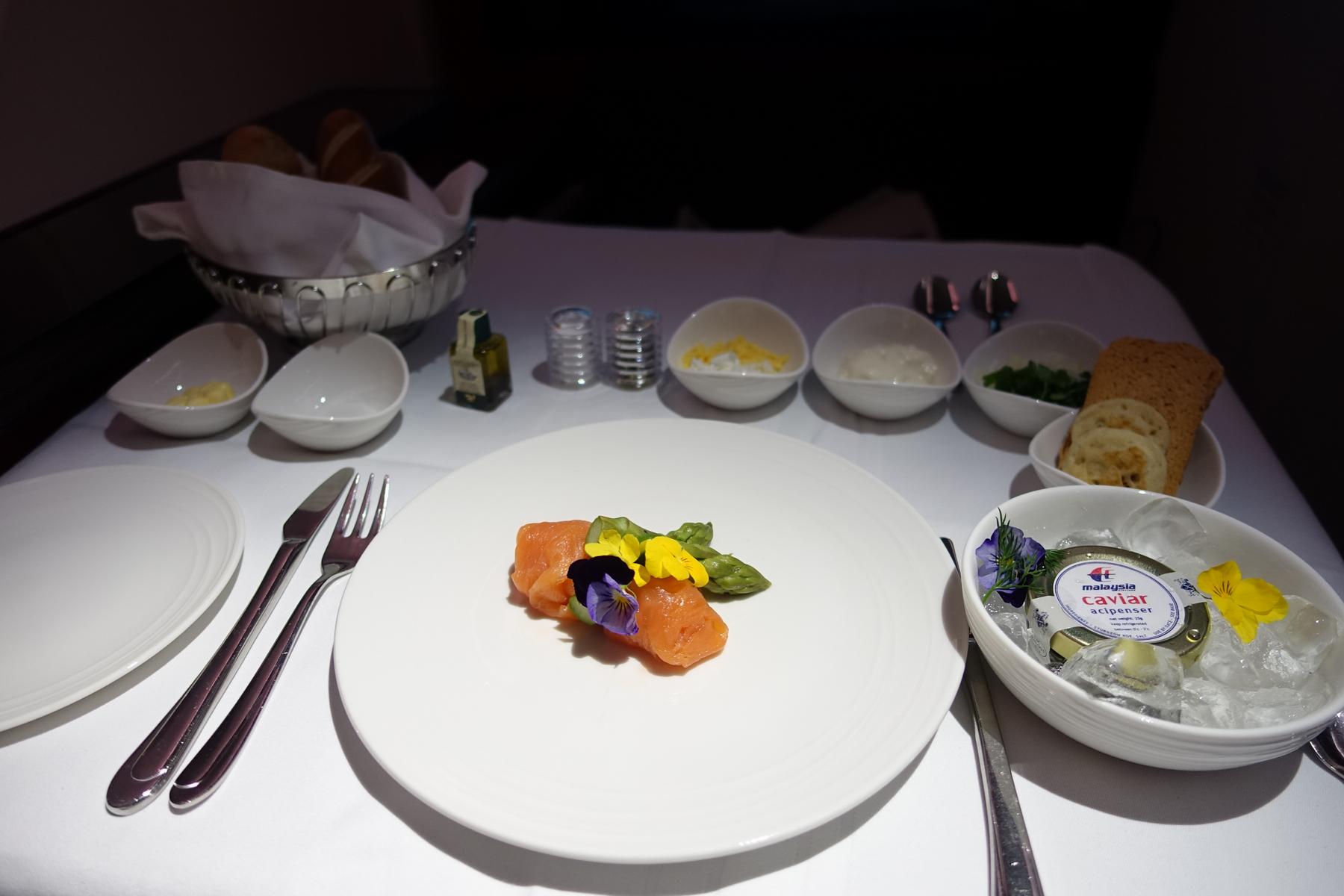 An elegantly plated Malaysia Airlines First Class meal featuring salmon, asparagus, caviar, and bread on a white table.