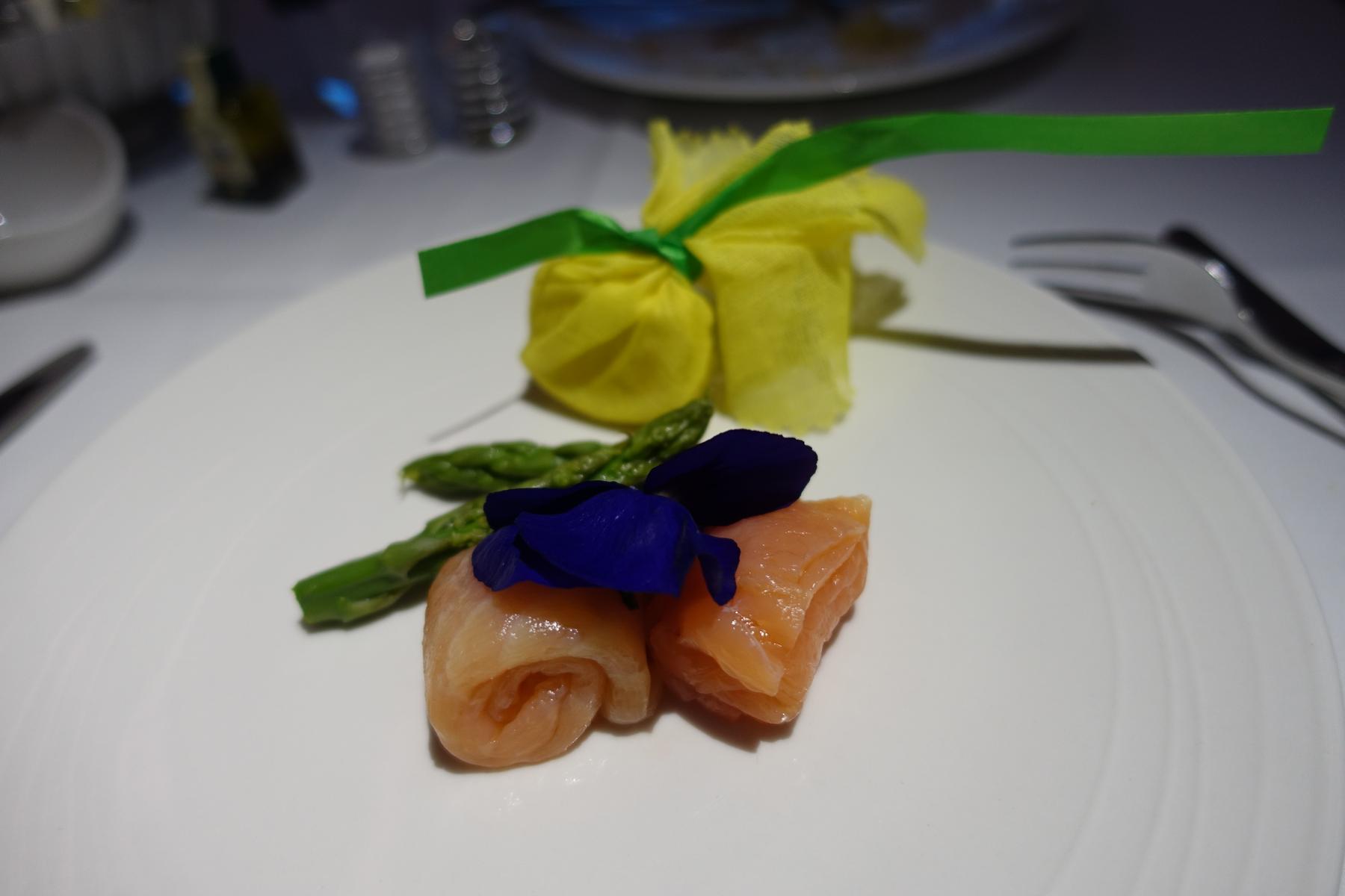 Salmon rolls topped with blue flowers, served with asparagus and yellow fabric parcels, on a white plate.