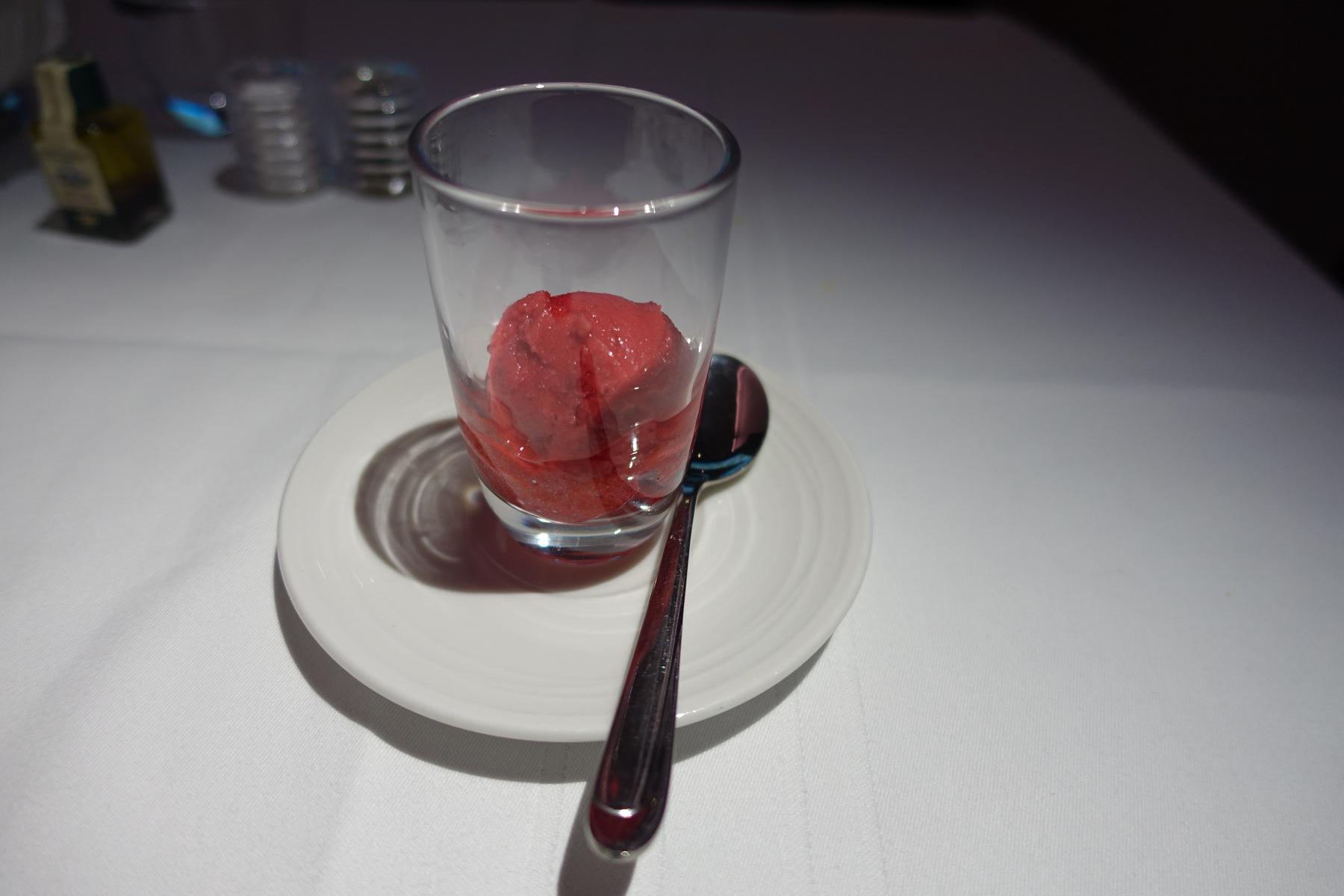 A glass of red sorbet with a spoon on a white table setting.