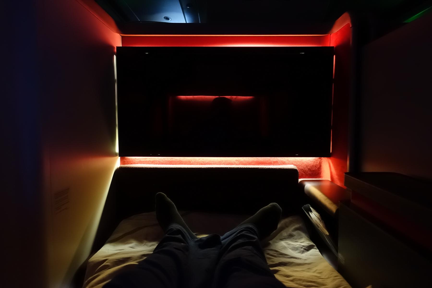 A passenger's feet are visible resting on a bed in a dimly lit Malaysia Airlines First Class suite with red accent lighting.