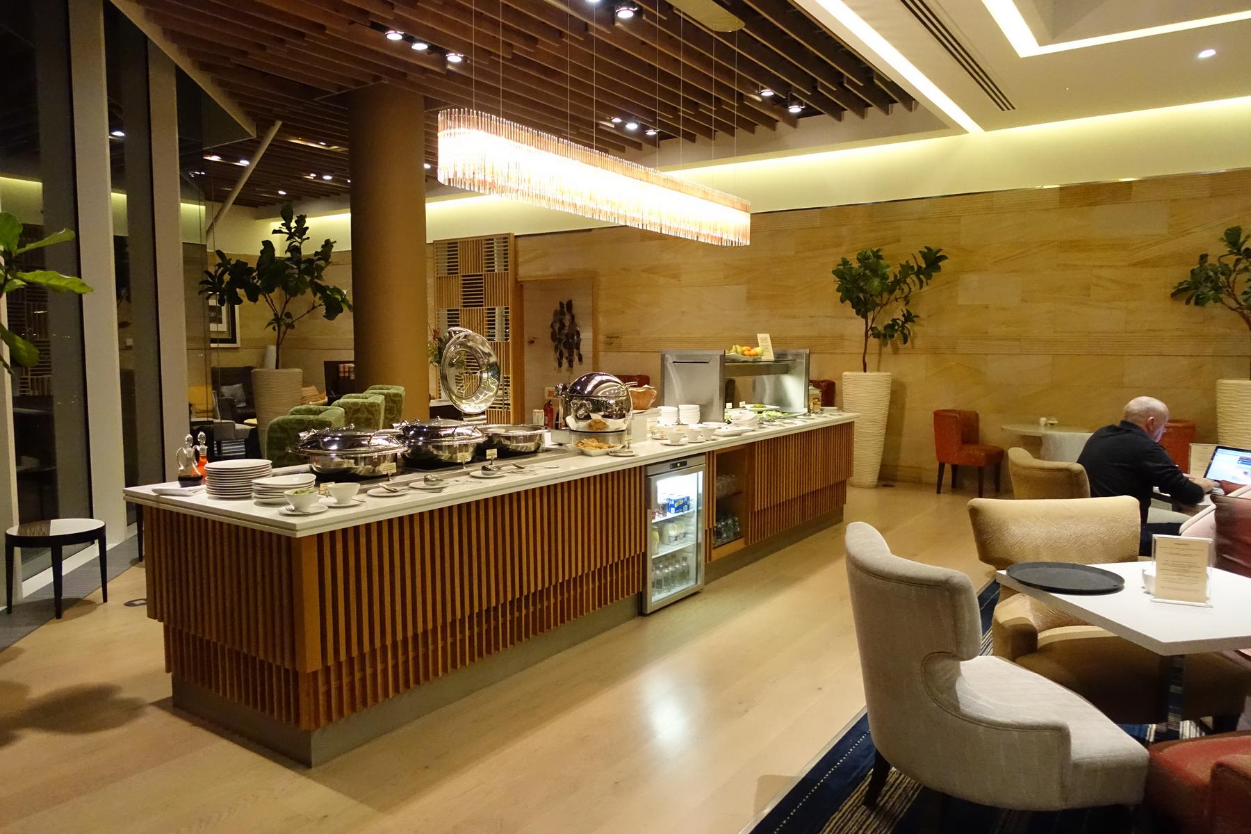 A long buffet counter filled with hot dishes and drinks stands in a modern, well-lit lounge.