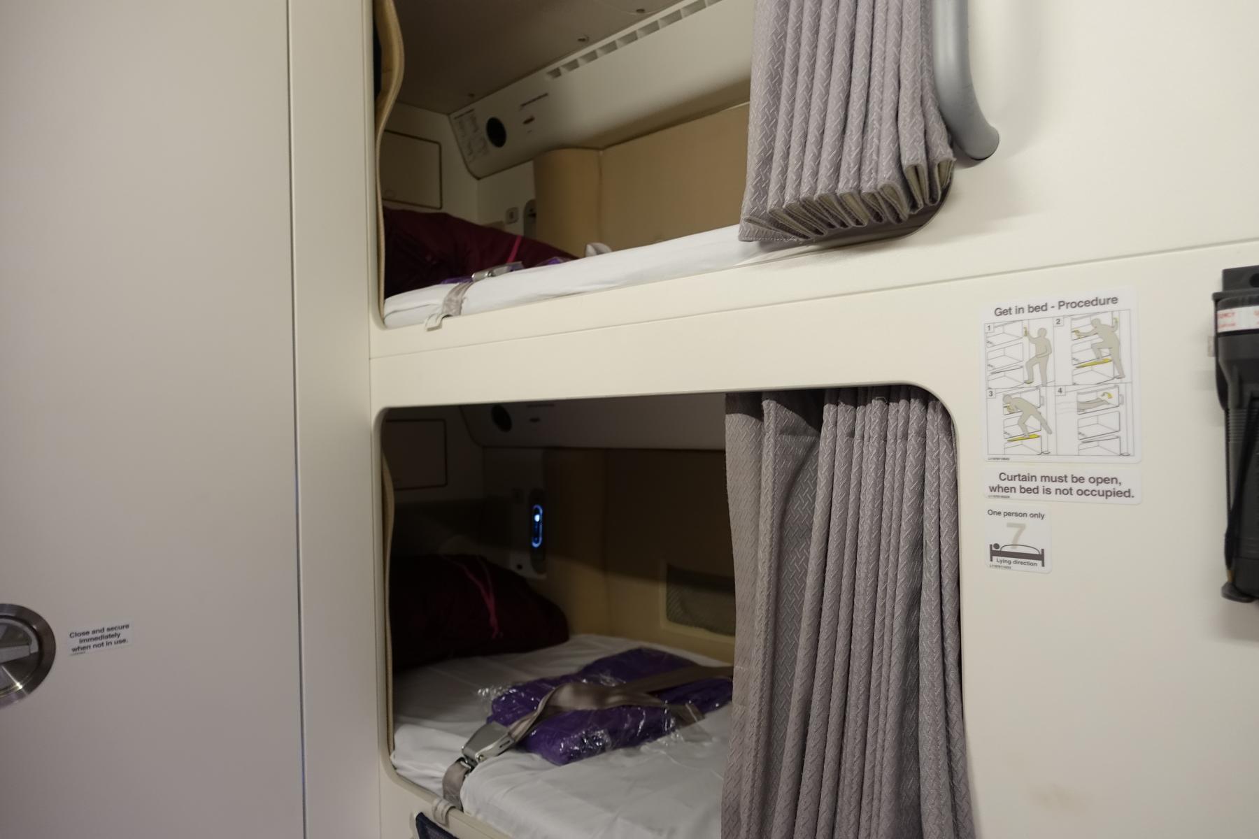 Two stacked aircraft bunk beds with seatbelts, privacy curtains, and a visible "Get in bed" procedure sign.