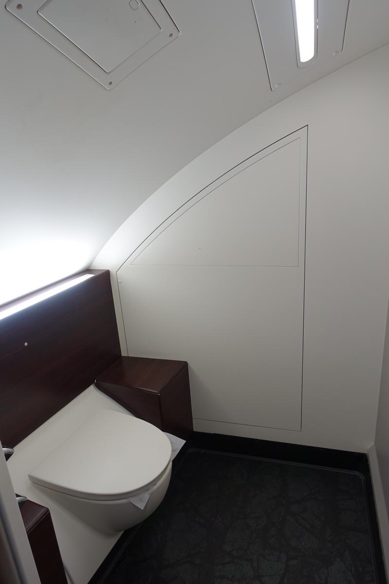 Interior of a clean, modern Malaysia Airlines First Class lavatory with a white toilet and dark wood details.