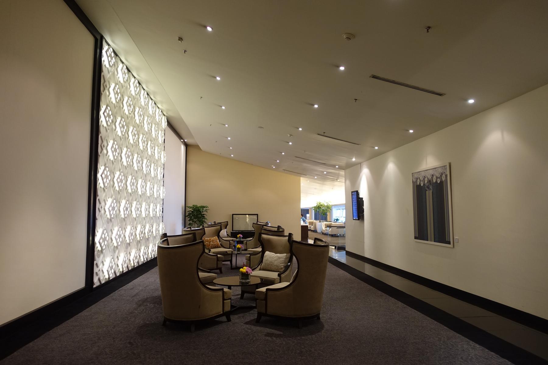 The Malaysian Airlines First Class Lounge features comfortable armchairs, small tables, and an illuminated patterned wall.