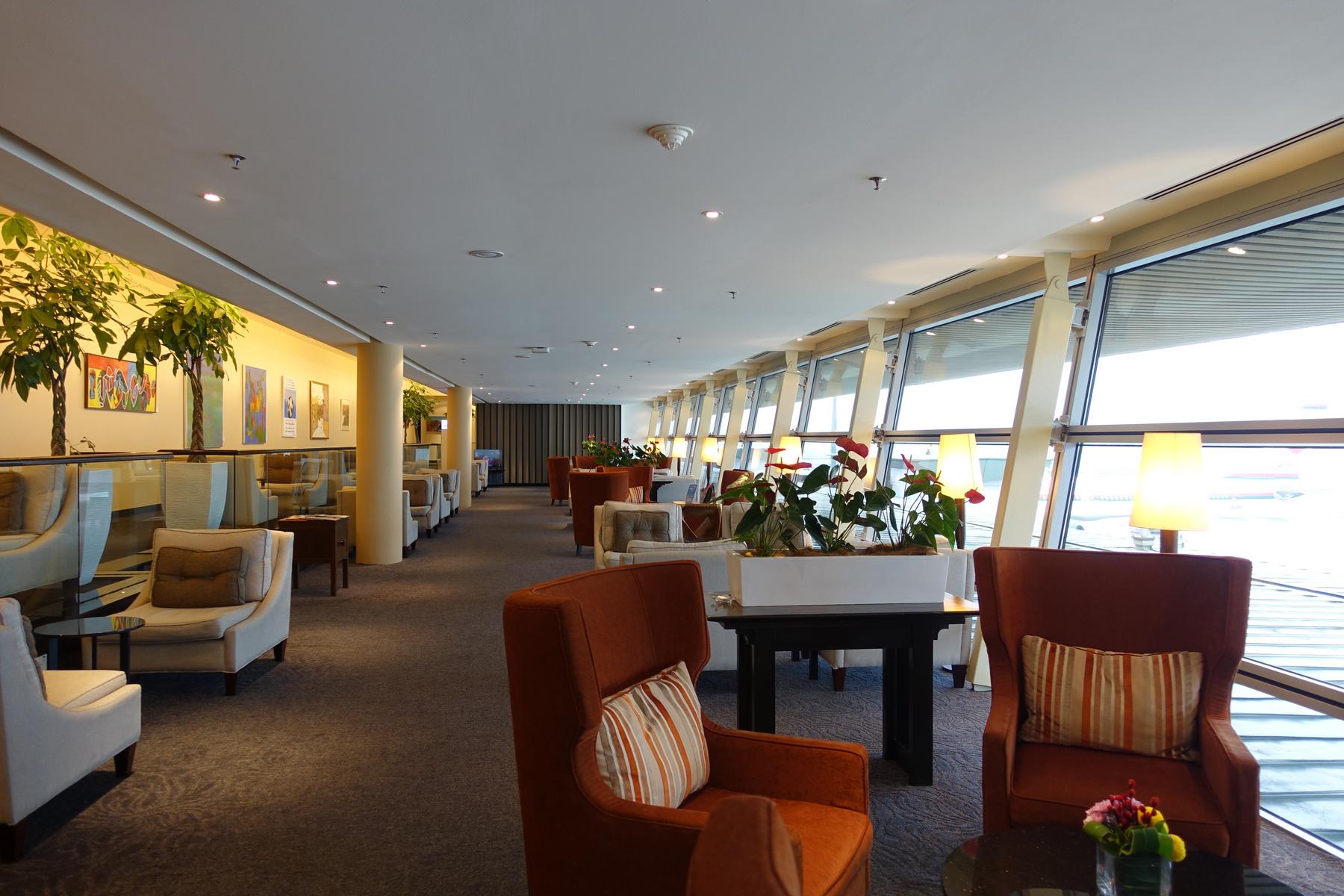 A spacious and brightly lit first-class airport lounge with comfortable seating and large windows overlooking the tarmac.
