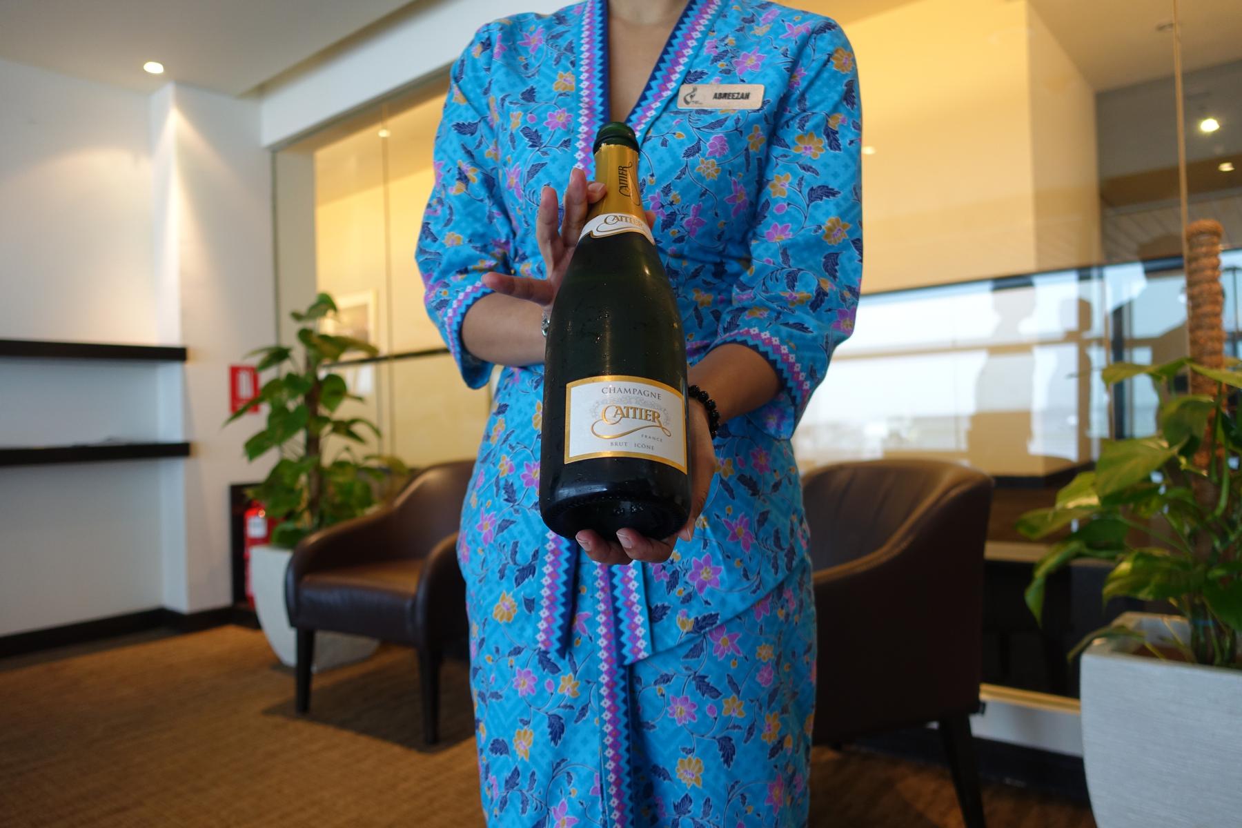 A Malaysian Airlines lounge attendant in a blue batik uniform holds a chilled bottle of Cattier Champagne.