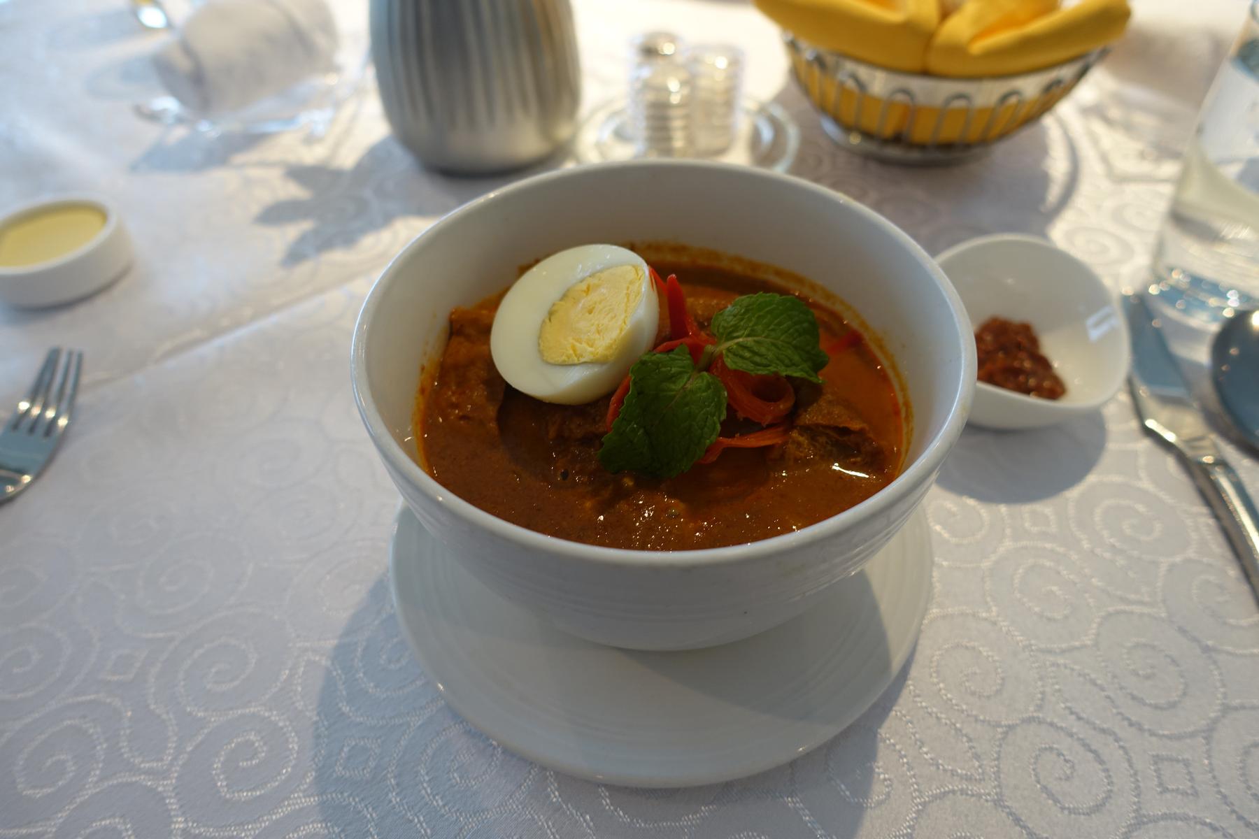 A bowl of Malaysian curry garnished with a hard-boiled egg and mint leaves, served on a white tablecloth.