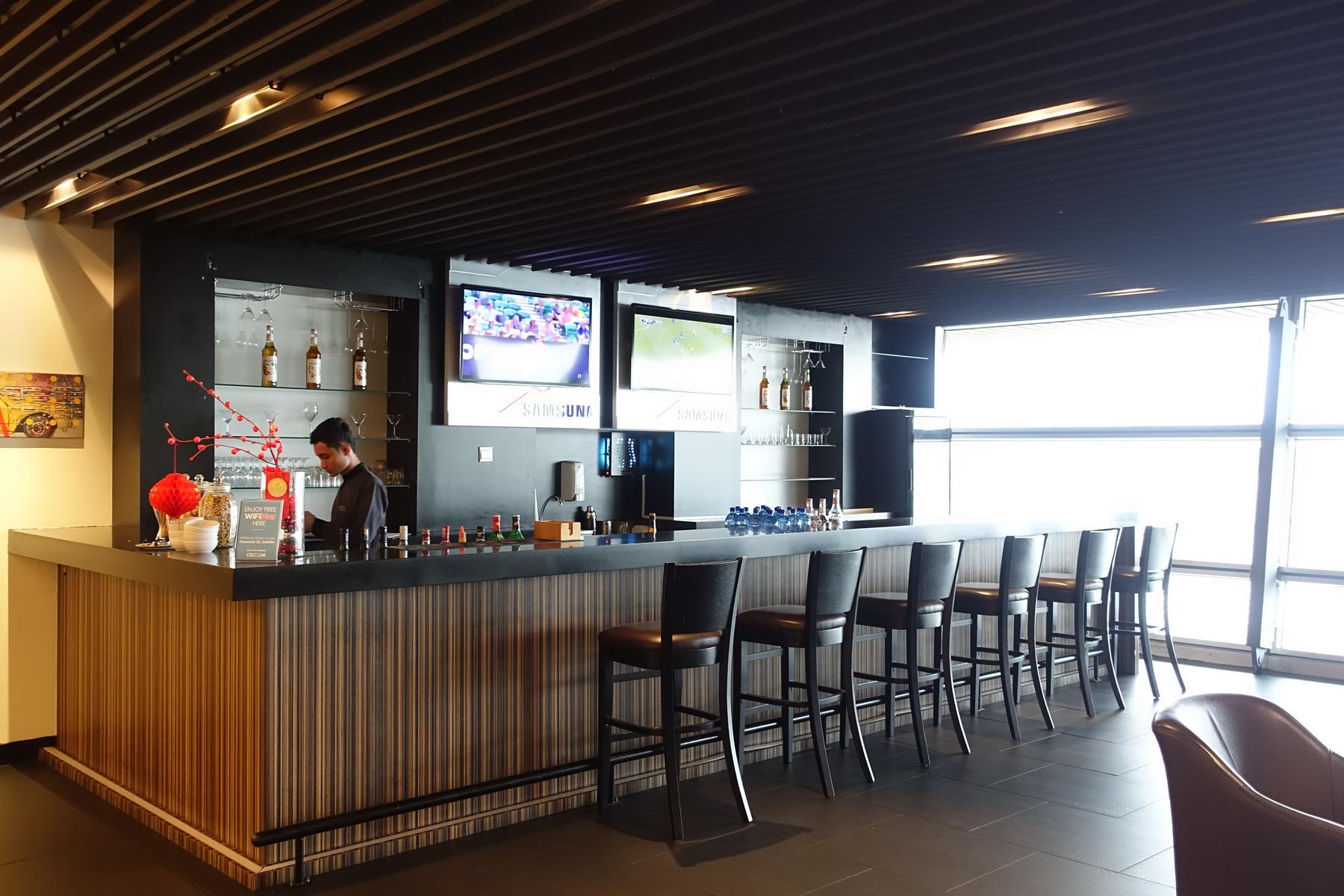 A modern bar area in a first-class airport lounge, with a bartender, stools, and large screens.