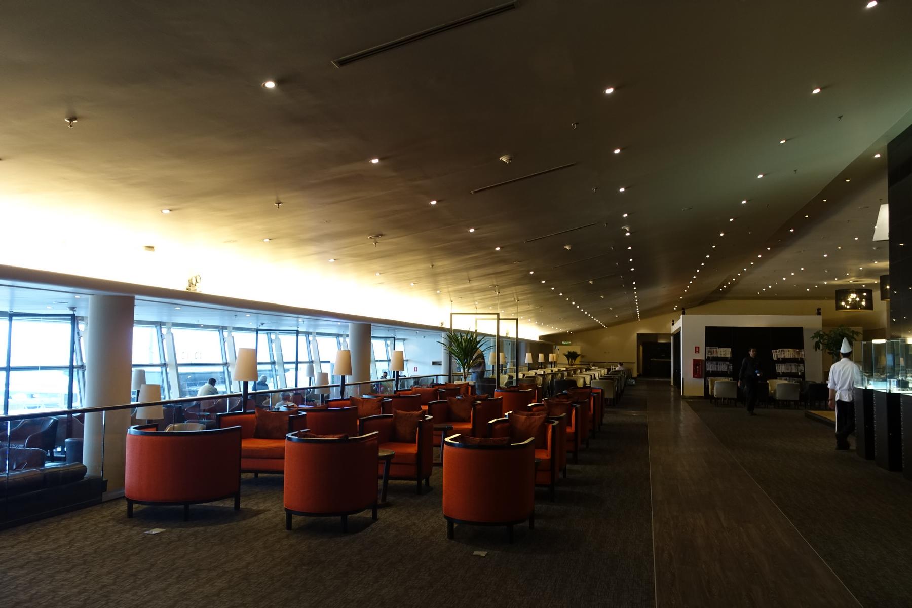 Spacious Malaysian Airlines First Class Lounge with orange seating areas, dining tables, and large windows.