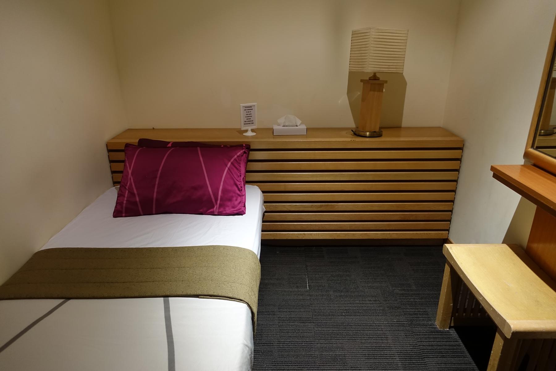 A small private room in an airport lounge featuring a bed, wooden headboard, and lamp.