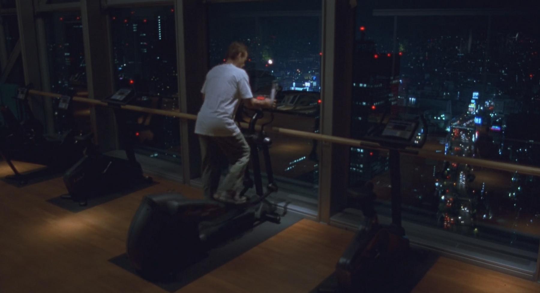 A person on an elliptical machine in a gym overlooking a brightly lit city at night.