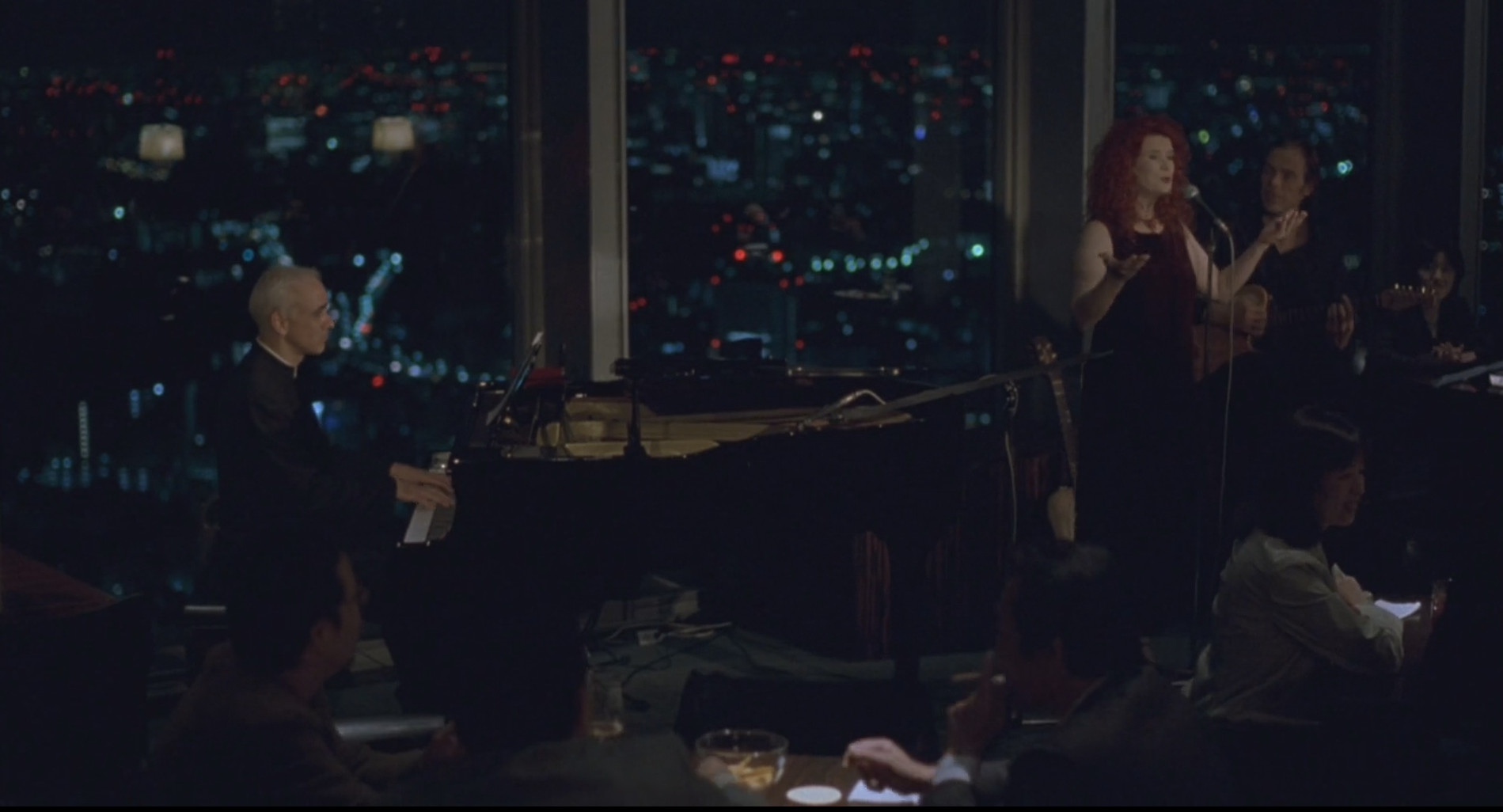 A singer and pianist perform live at the dimly lit New York Bar, with blurred city lights visible at night.