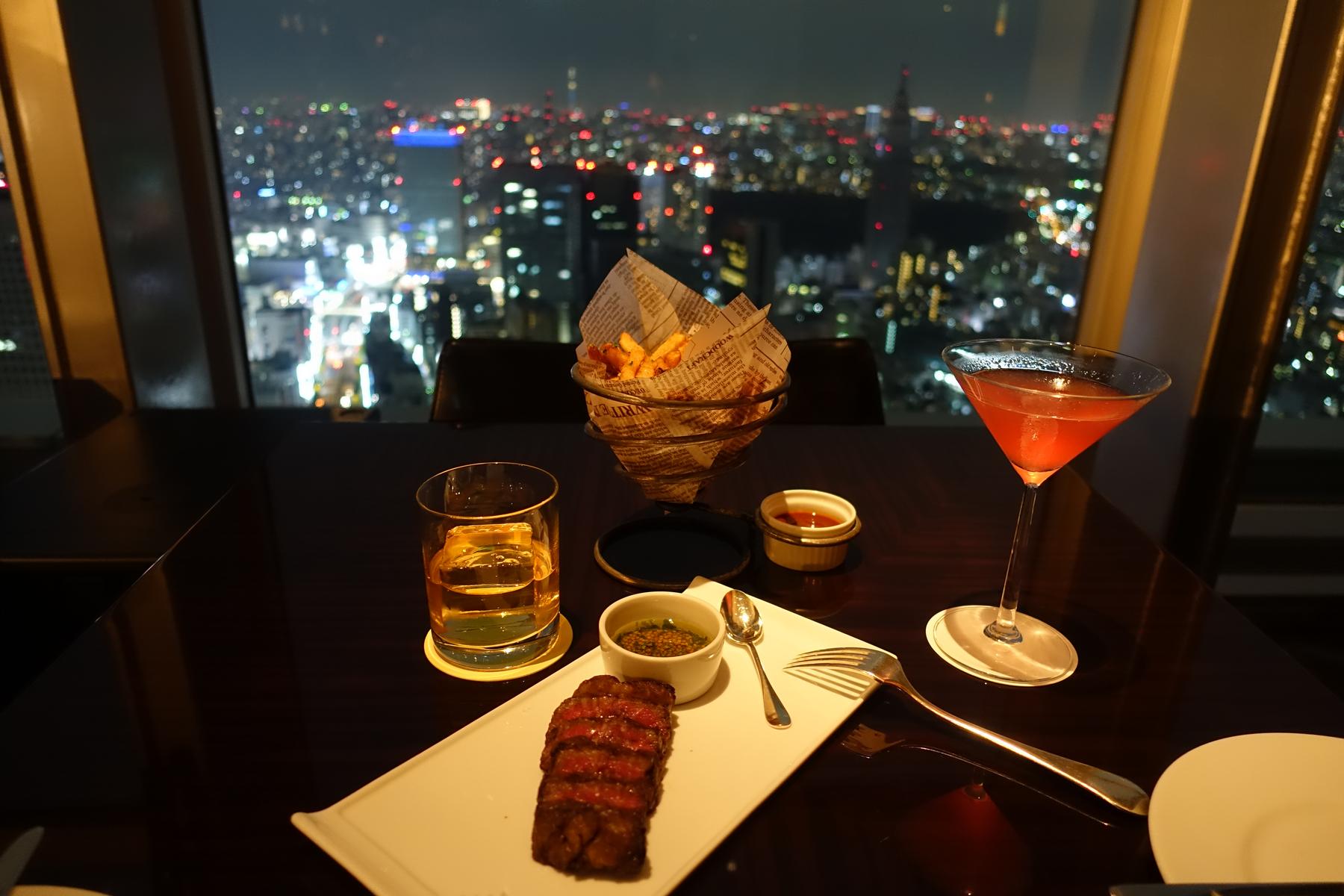 A table with sliced steak, fries, and drinks overlooks a sparkling city skyline at night.