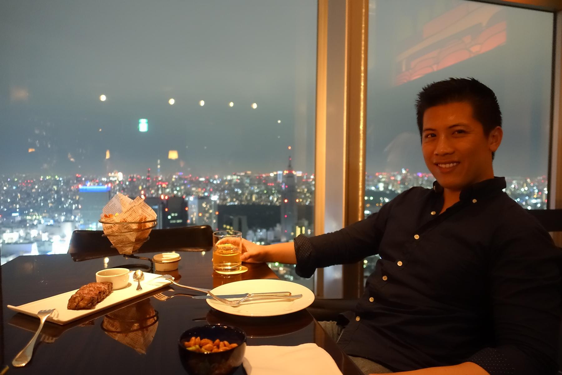 A man smiles at the Park Hyatt Tokyo's New York Bar, enjoying a drink and food with a night view of the city skyline.