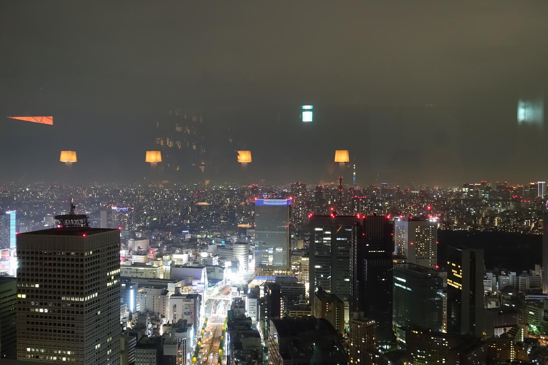 Nighttime cityscape from a high window, with reflections of indoor lamps above the brightly lit buildings.