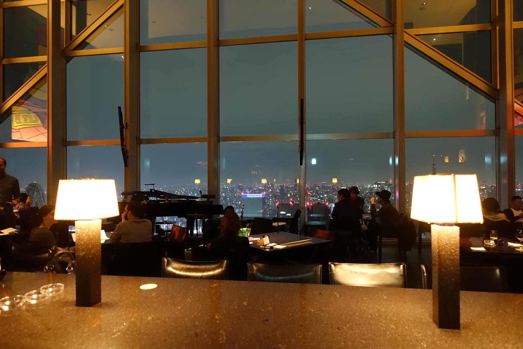Guests relax in a dimly lit bar overlooking a Tokyo city skyline at night, with a piano and glowing lamps in the foreground.