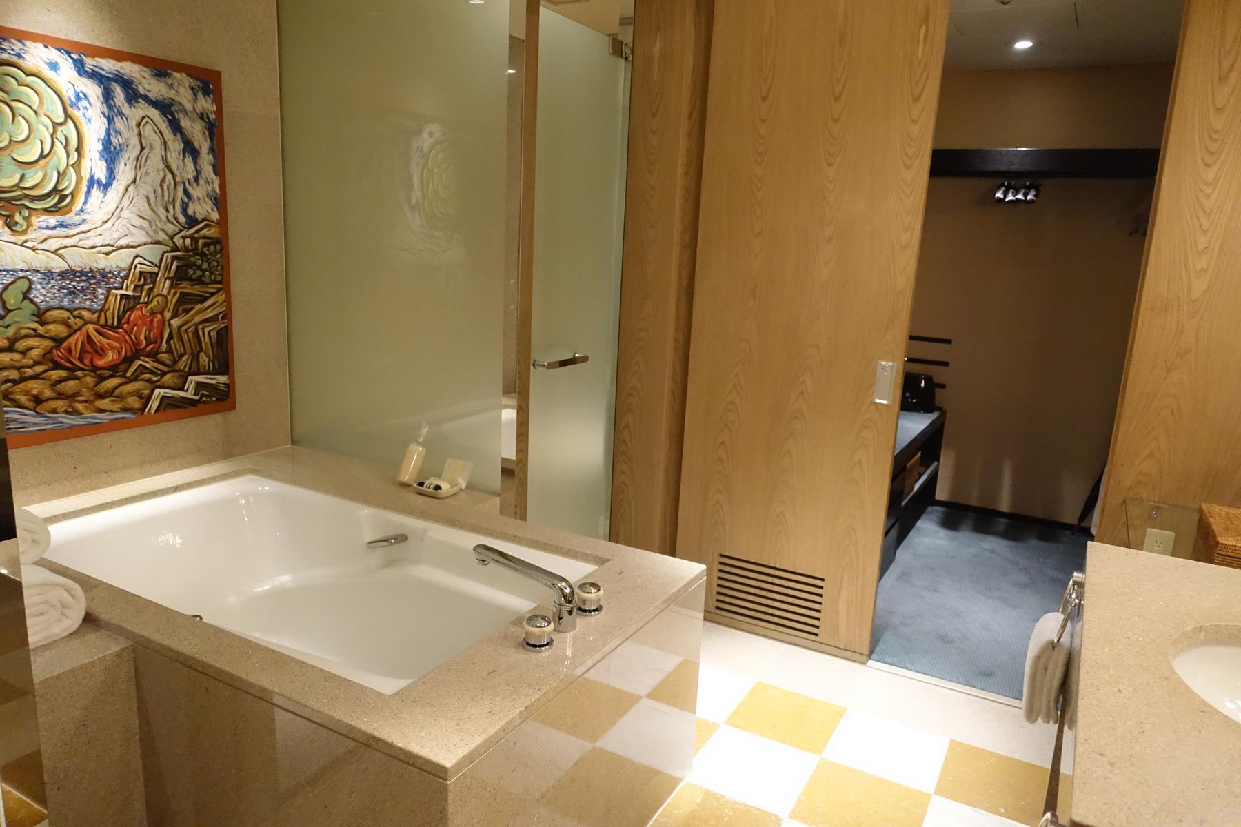 A luxurious hotel bathroom featuring a large bathtub, vibrant artwork, and an open doorway to a dressing area.