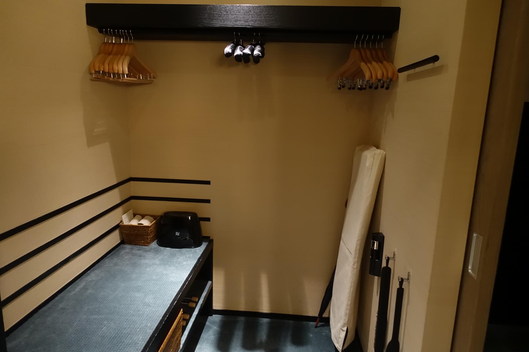 A hotel closet area featuring wooden hangers, a bench with slippers and a black amenity, and a futon mattress leaning against a beige wall.