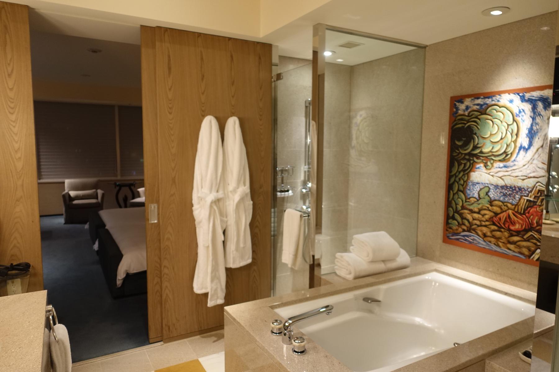 An elegant hotel bathroom with a large bathtub, two white bathrobes, a colorful painting, and a view into an adjacent bedroom.