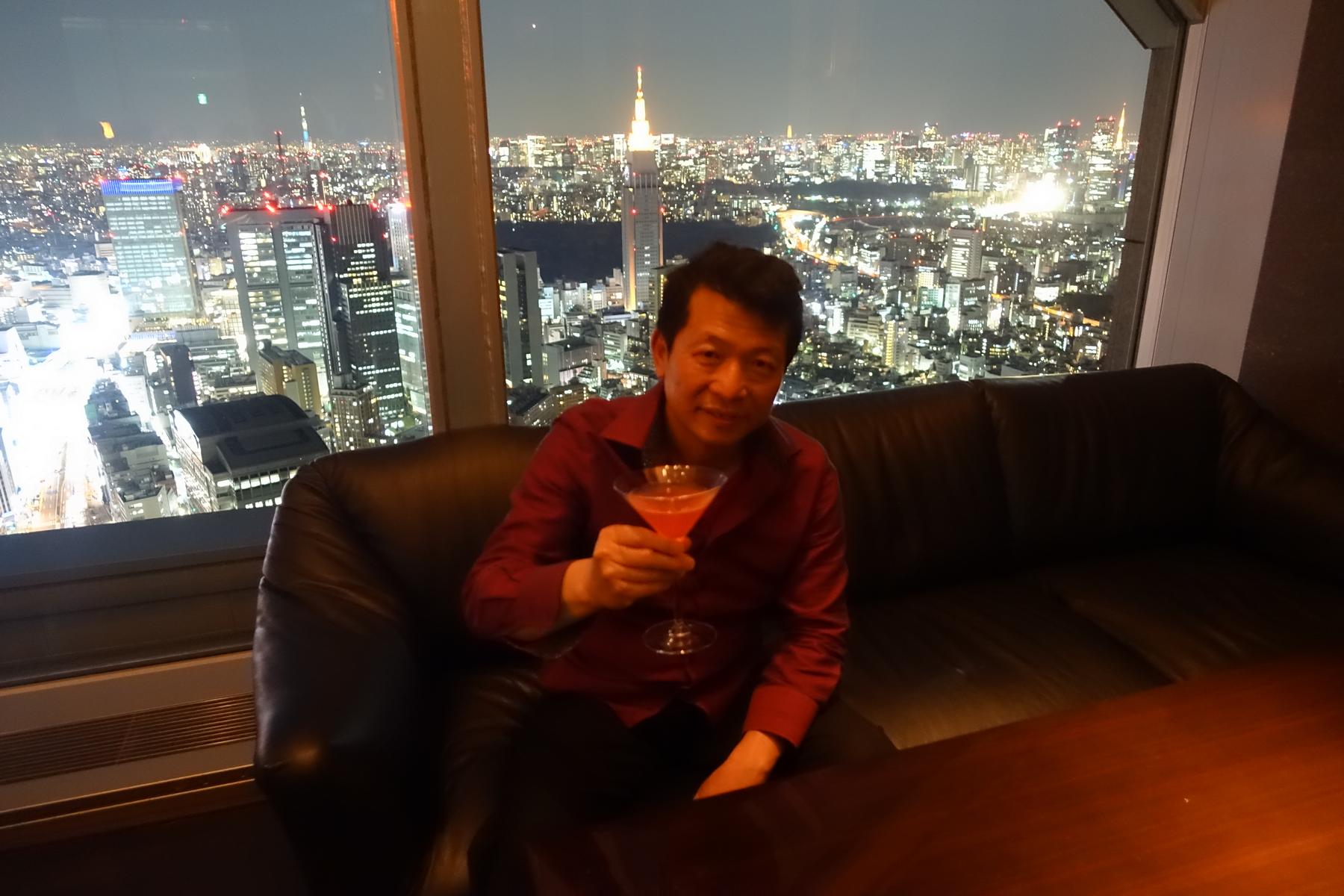 A man in a red shirt holds a cocktail against a backdrop of Tokyo's night skyline.