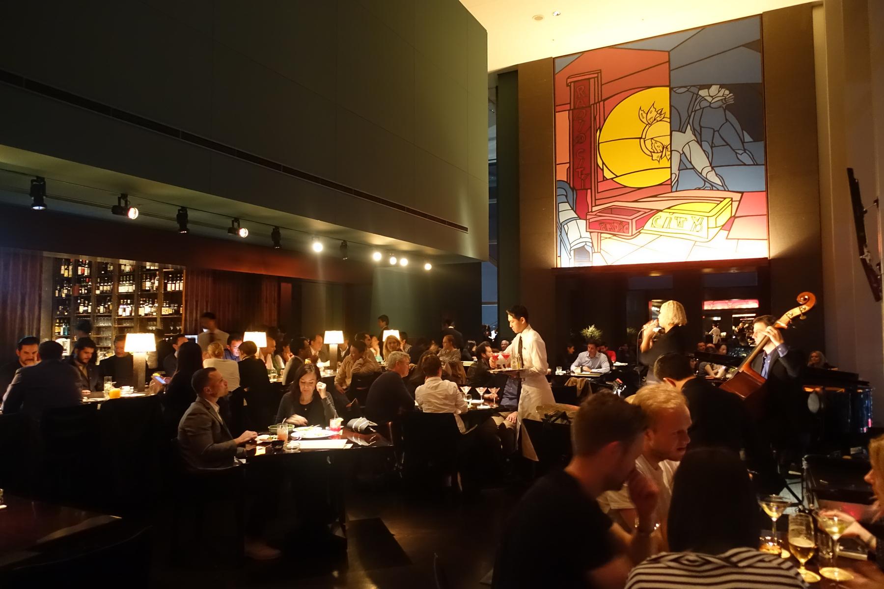 The Park Hyatt Tokyo's New York Bar is filled with guests, a server, live jazz music, and features a large colorful mural.
