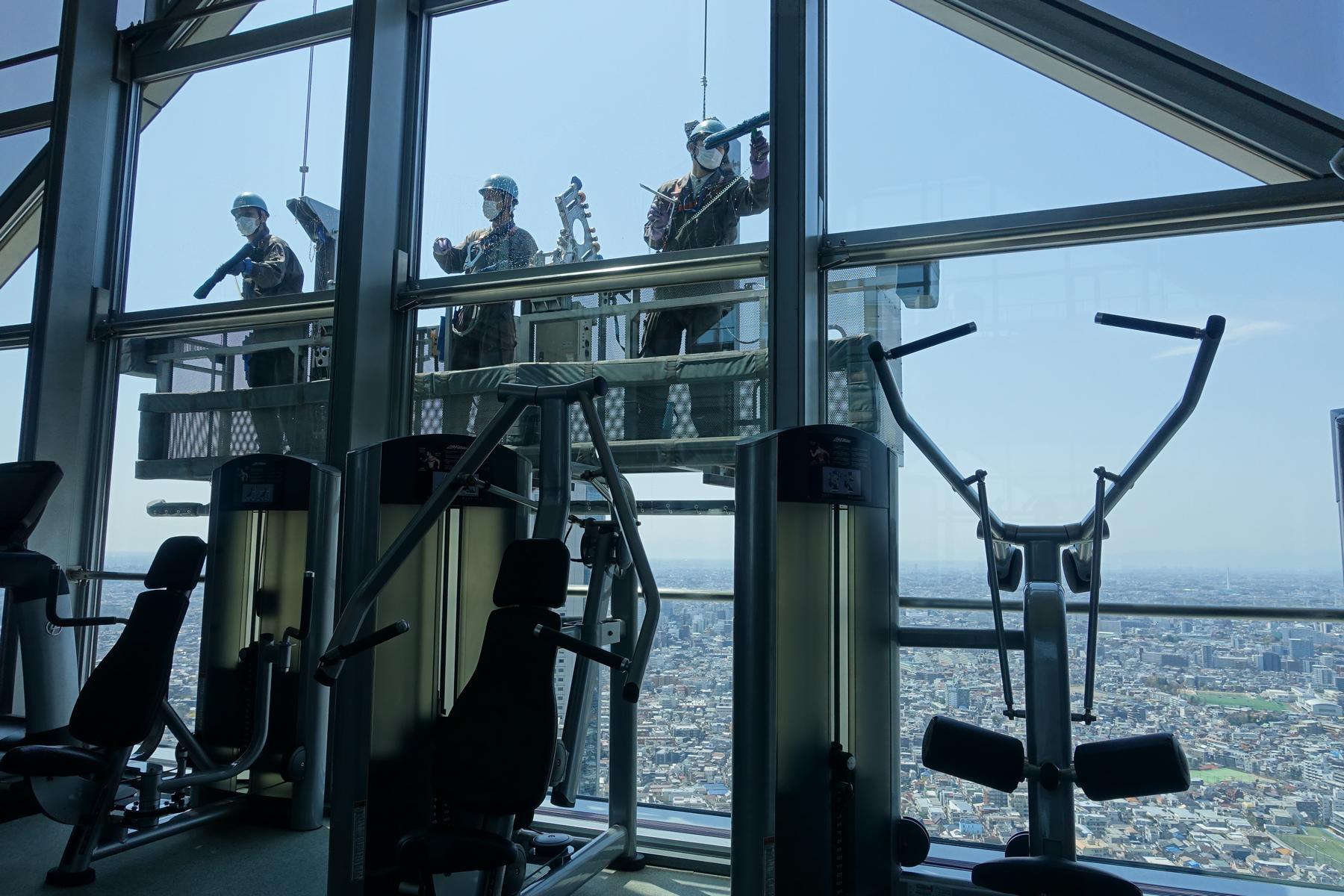 Window cleaners on a platform outside a high-rise gym with city views.