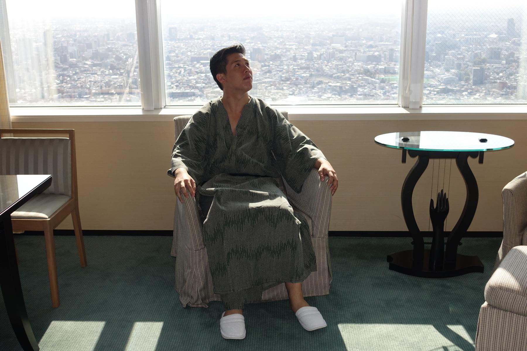 A man in a dark green patterned yukata and white slippers sits in an armchair, looking up at the city view from a high-rise window.