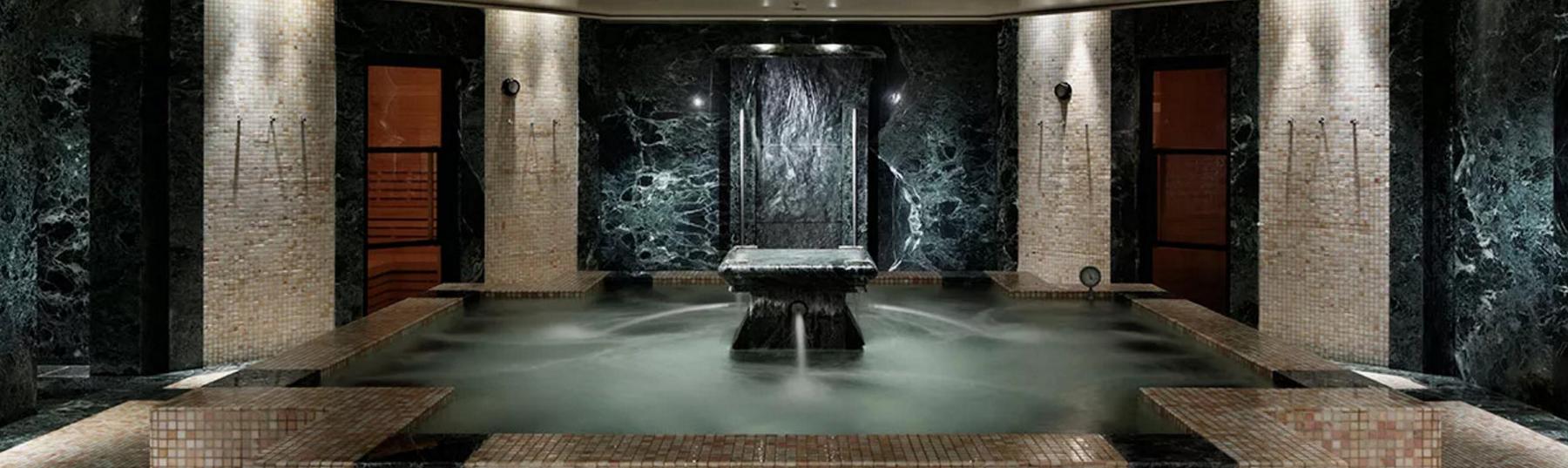 Indoor spa with a large pool and central water feature, surrounded by dark green marble and light mosaic walls.