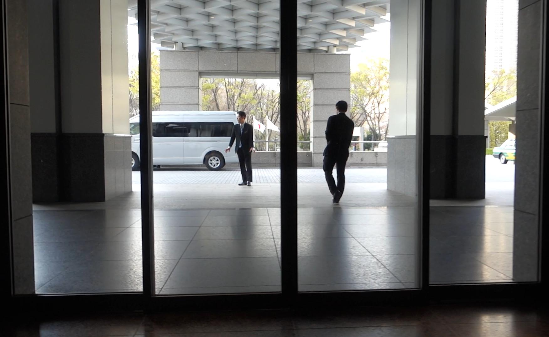 A view through glass doors shows two men and a white van at the hotel entrance.