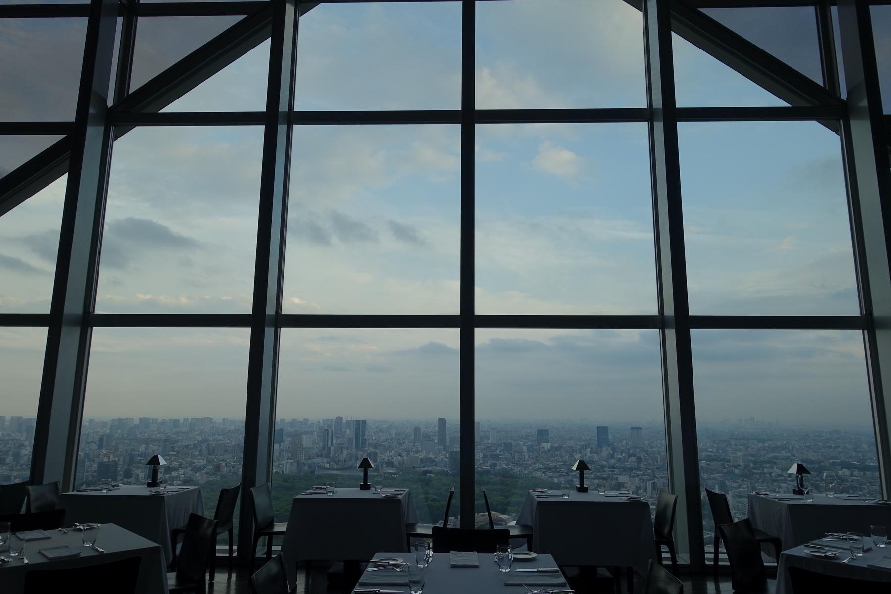 A high-rise restaurant with large grid-pattern windows overlooks a sprawling city skyline under a cloudy sky, with dining tables set in the foreground.