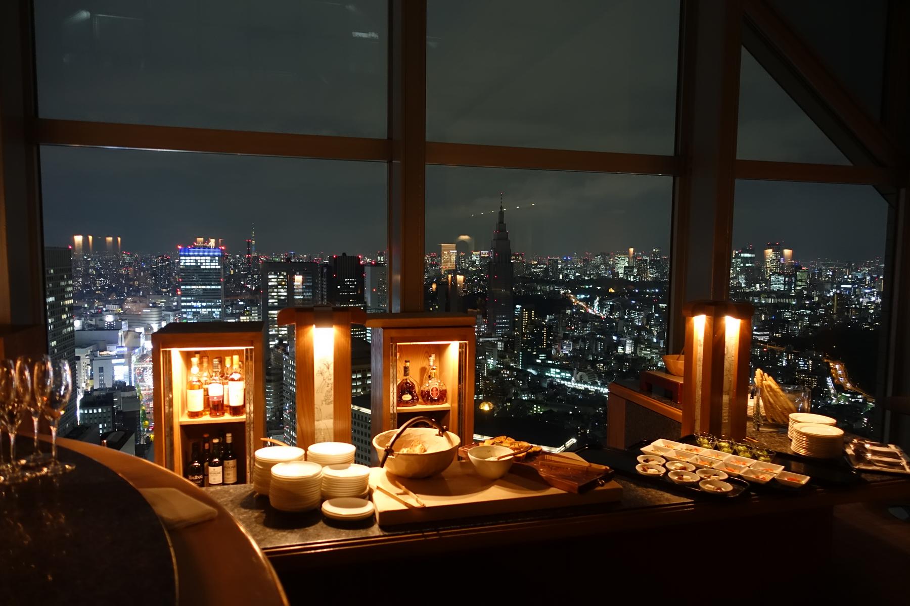 A bar counter with glowing displays of bottles and food overlooks a vast, illuminated Tokyo cityscape at night.