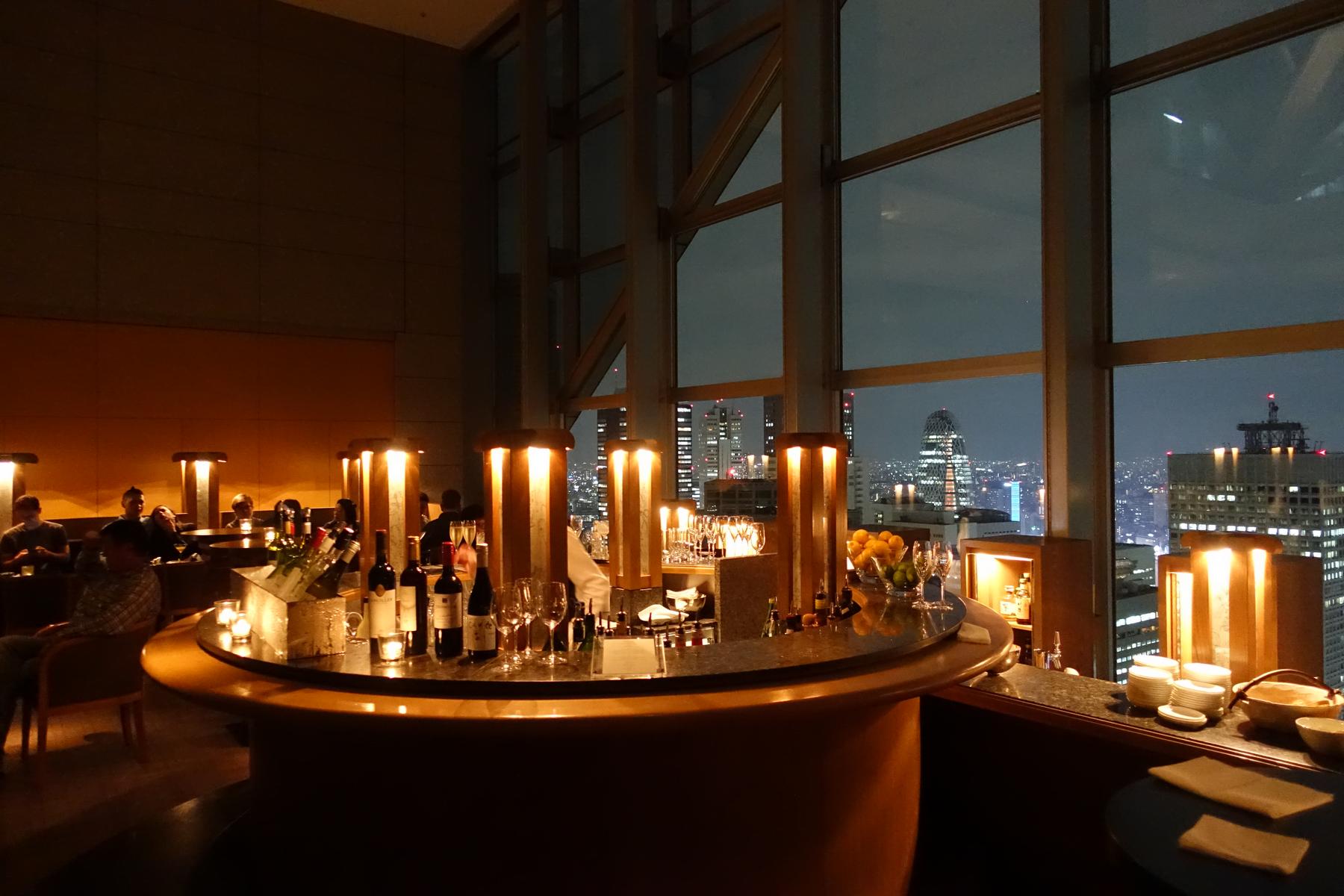 A dimly lit bar with a circular counter and large windows offering a night view of a city skyline.