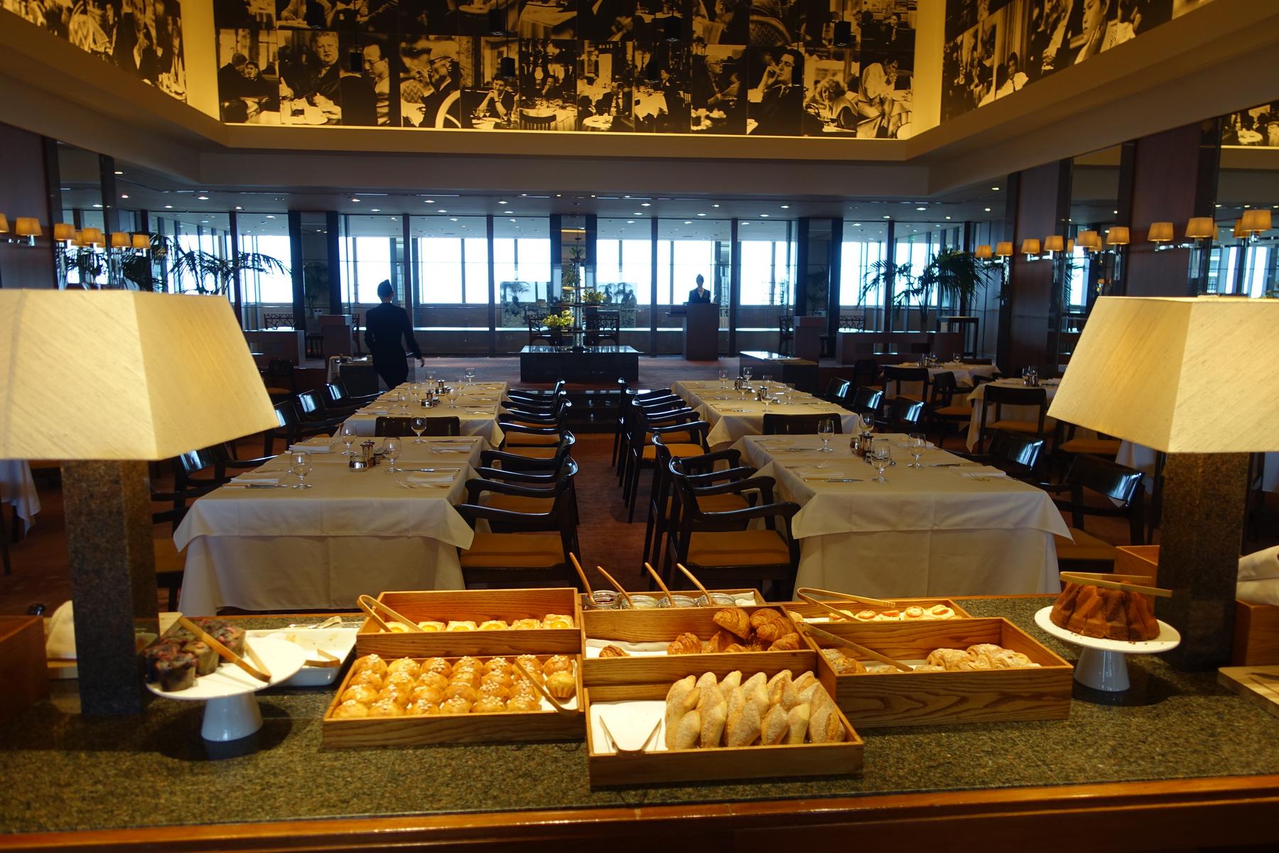 A breakfast buffet with pastries and bread in the foreground, with set dining tables and a spacious dining room behind.
