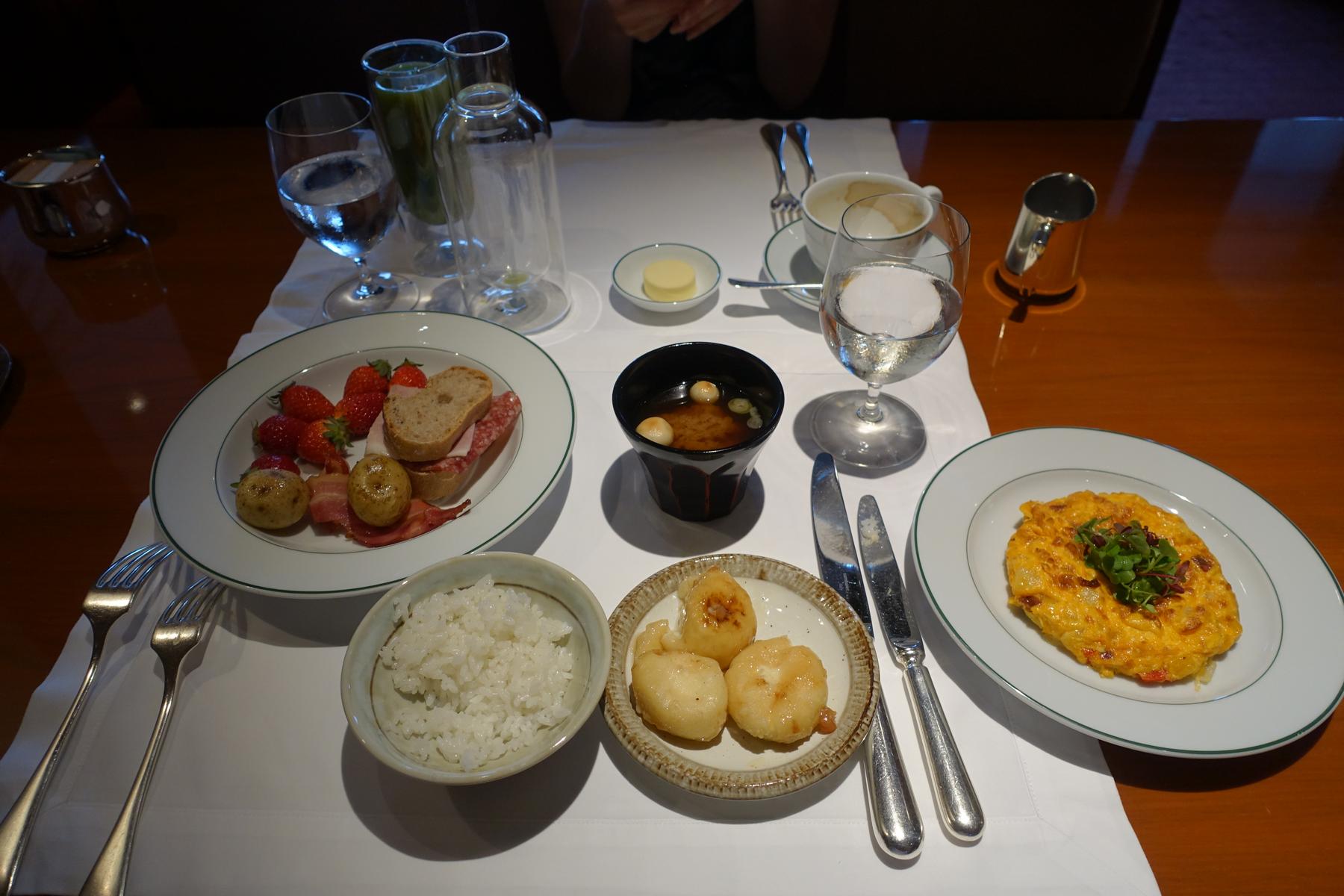 A diverse breakfast includes an omelet, fresh fruit, bacon, bread, rice, and soup arranged on a white tablecloth.