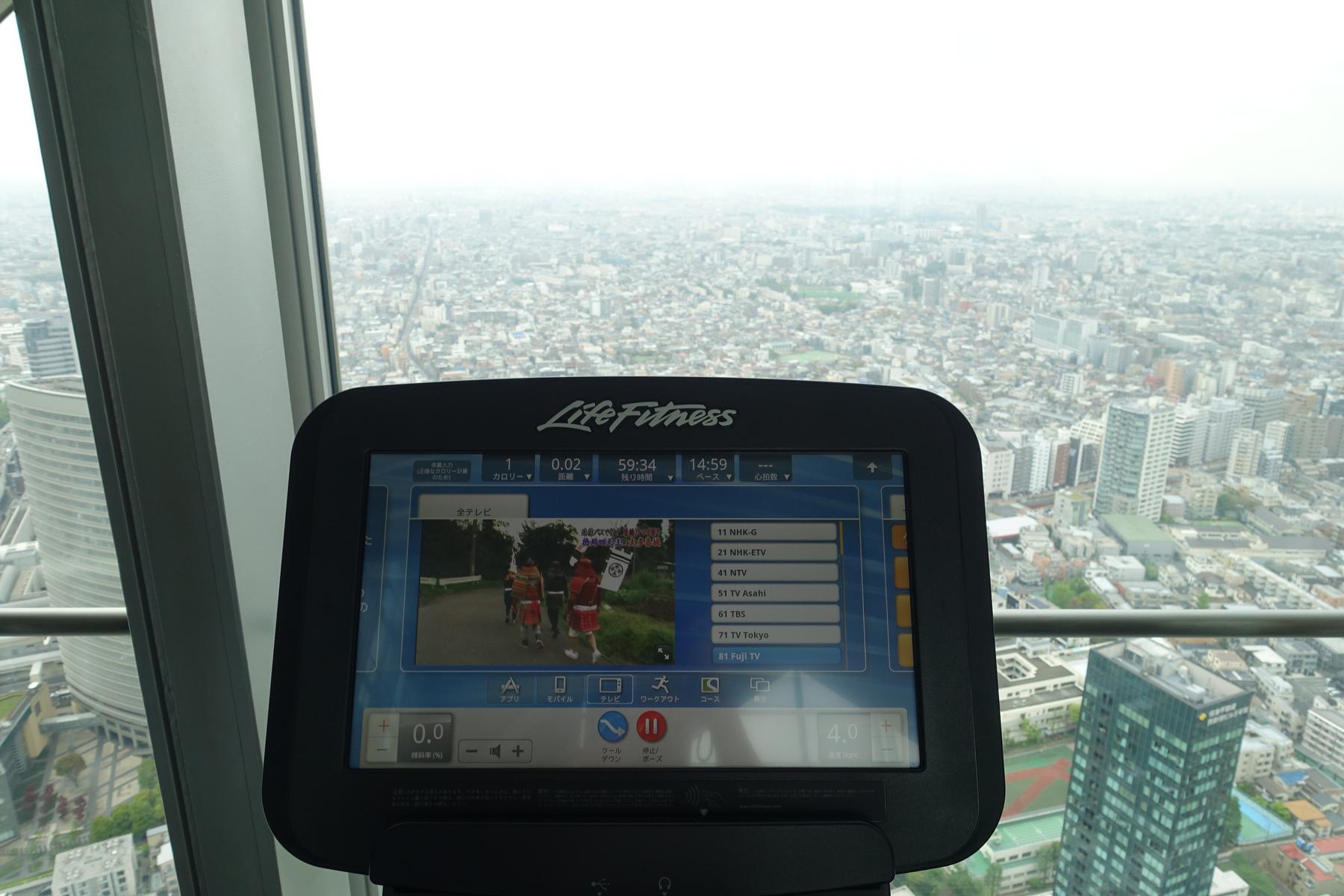A Life Fitness exercise machine screen displays TV channels and workout stats, with a panoramic view of the Tokyo cityscape from a high floor.