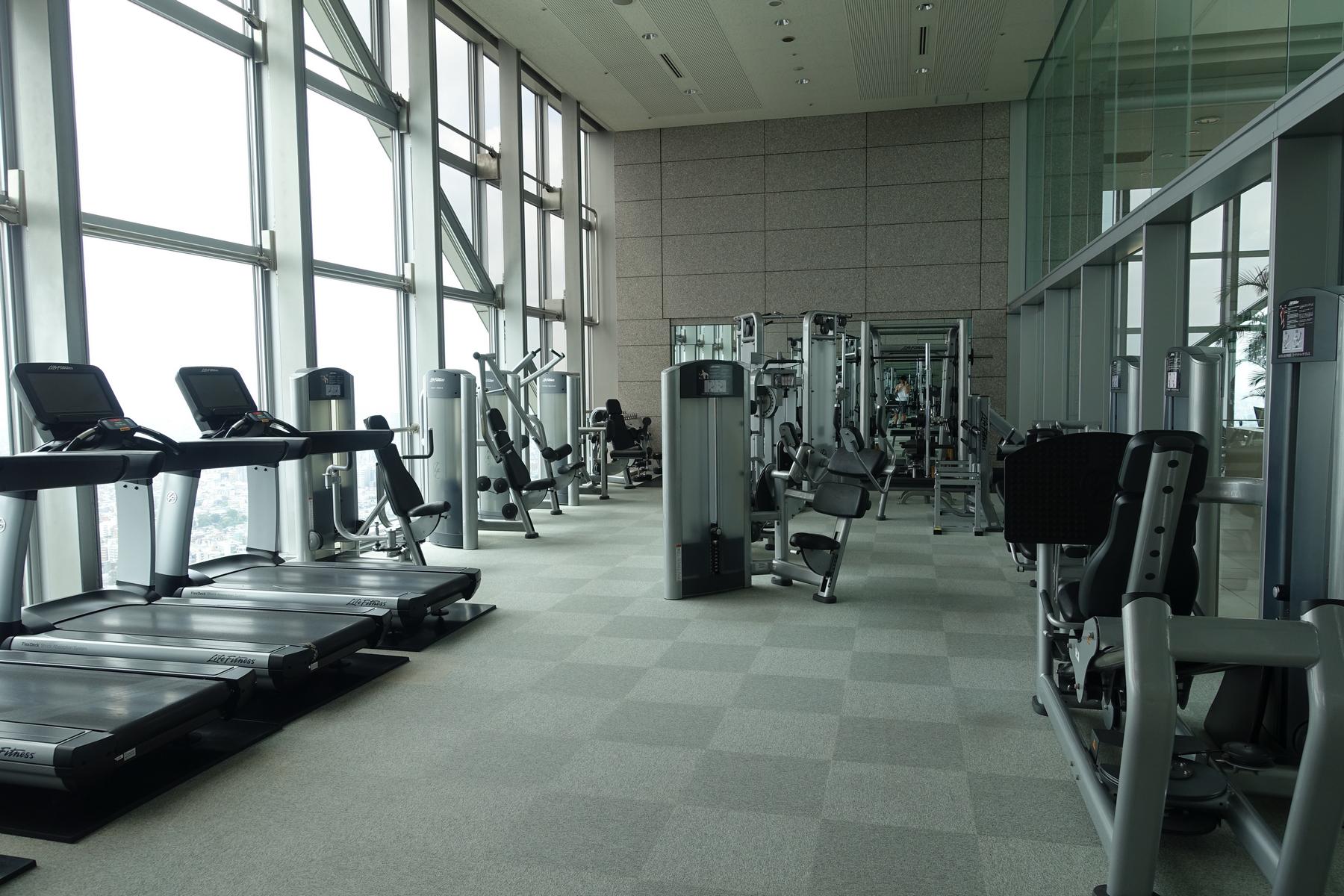 A spacious, modern gym with various exercise machines and large windows offering city views.